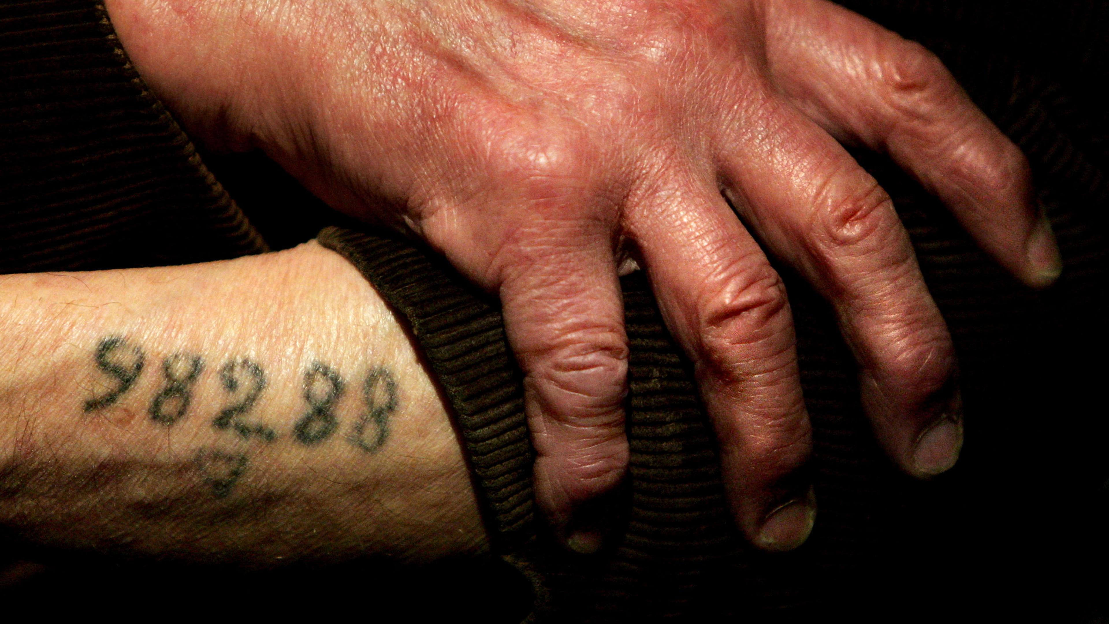 Photo of an elderly hand resting on a wrist, tattooed with the number 58288, on a dark fabric background.