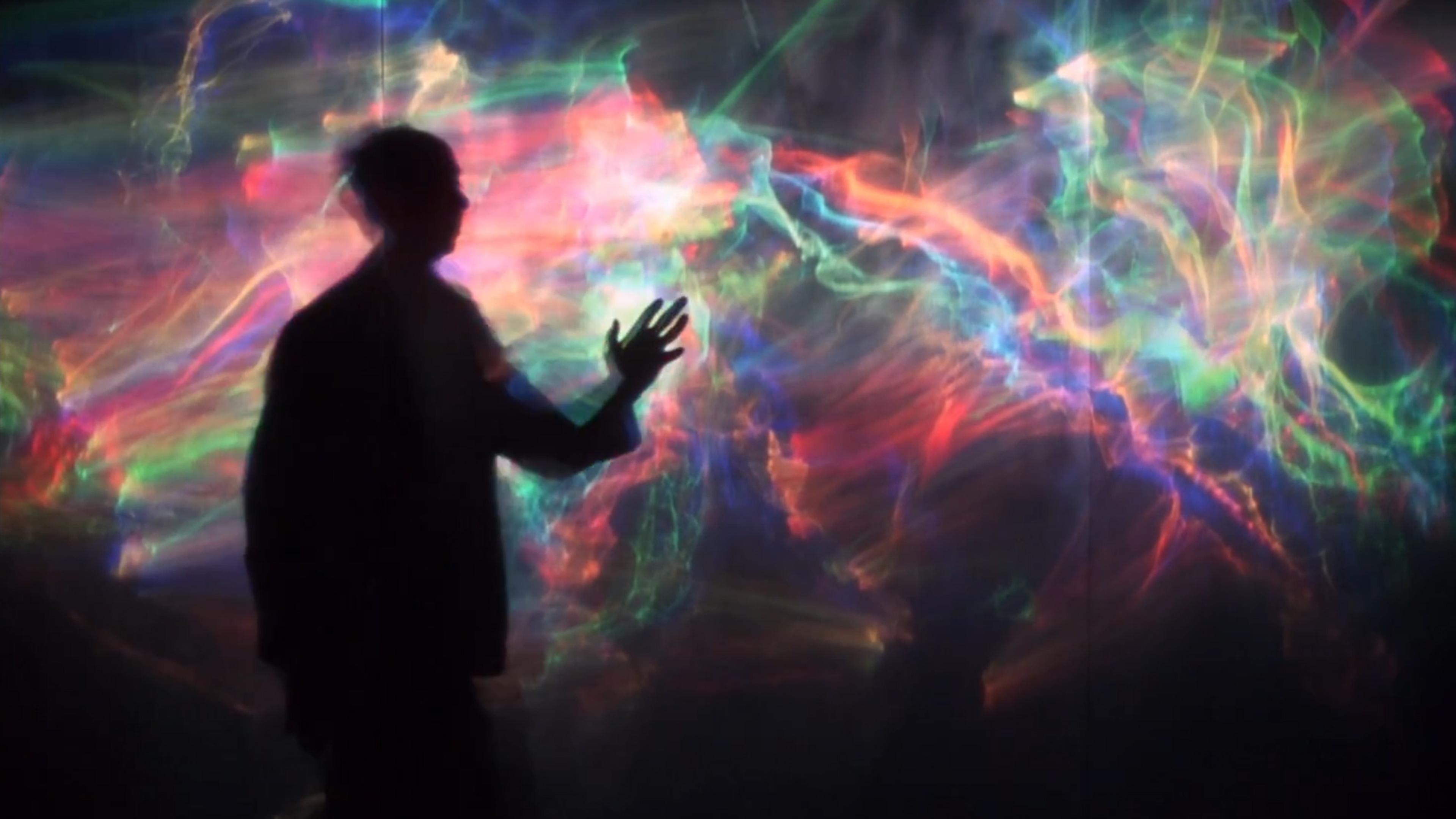 Silhouette of person reaching towards vibrant colourful light patterns on a wall.