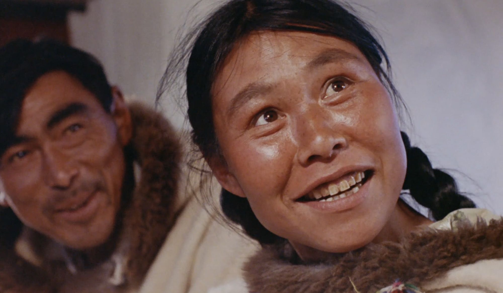 A film still showing the Inuit artist Kenojuak Ashevak and her husband Johnniebo in fur-trimmed clothing, smiling and looking off camera.