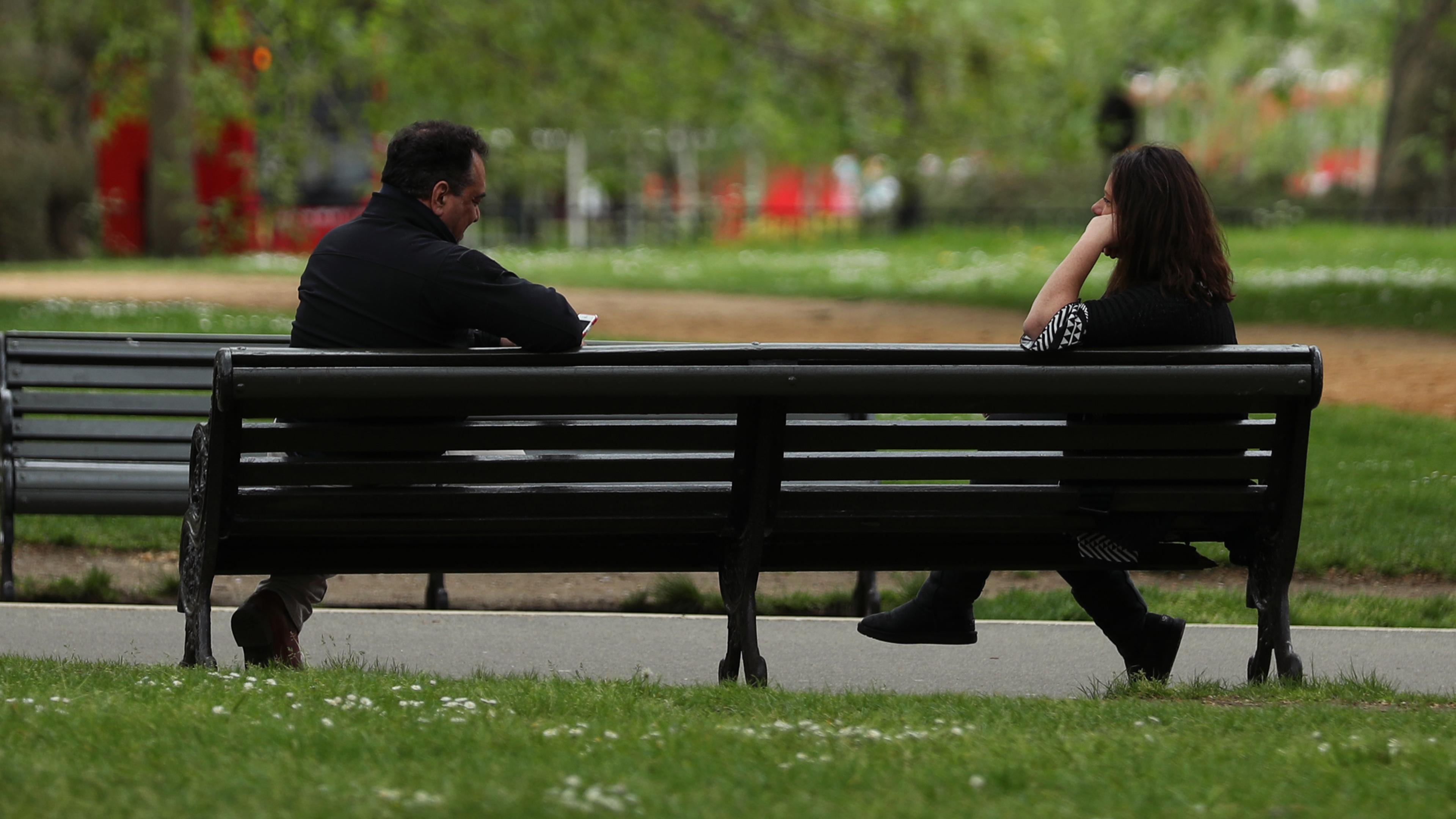 Photo of two people sitting on a park bench in a lush green setting with a pathway and trees in the background.