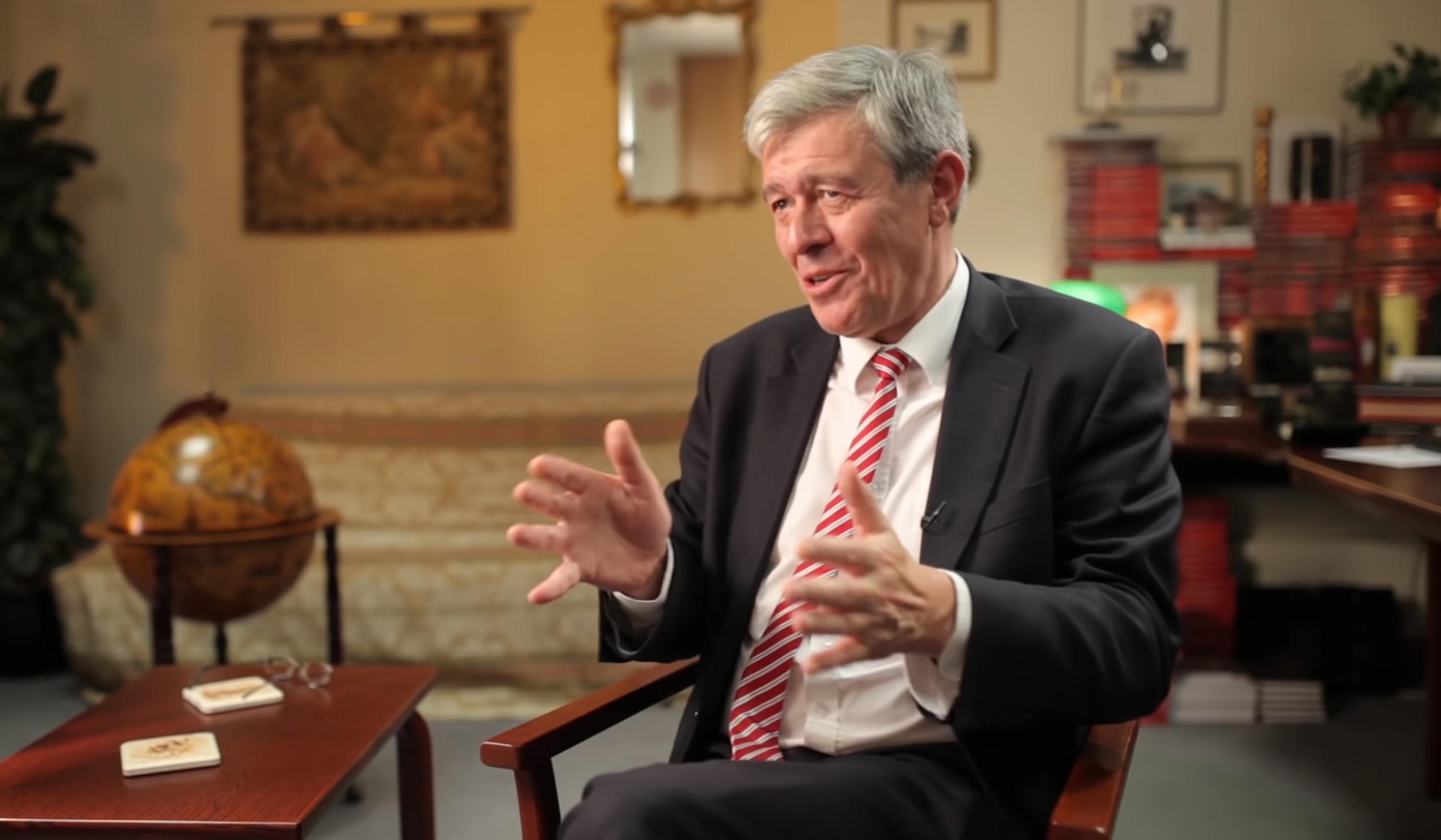 An older man in a suit and striped tie gestures while speaking in a cosy, book-filled office with a globe and a decorated wall hanging.