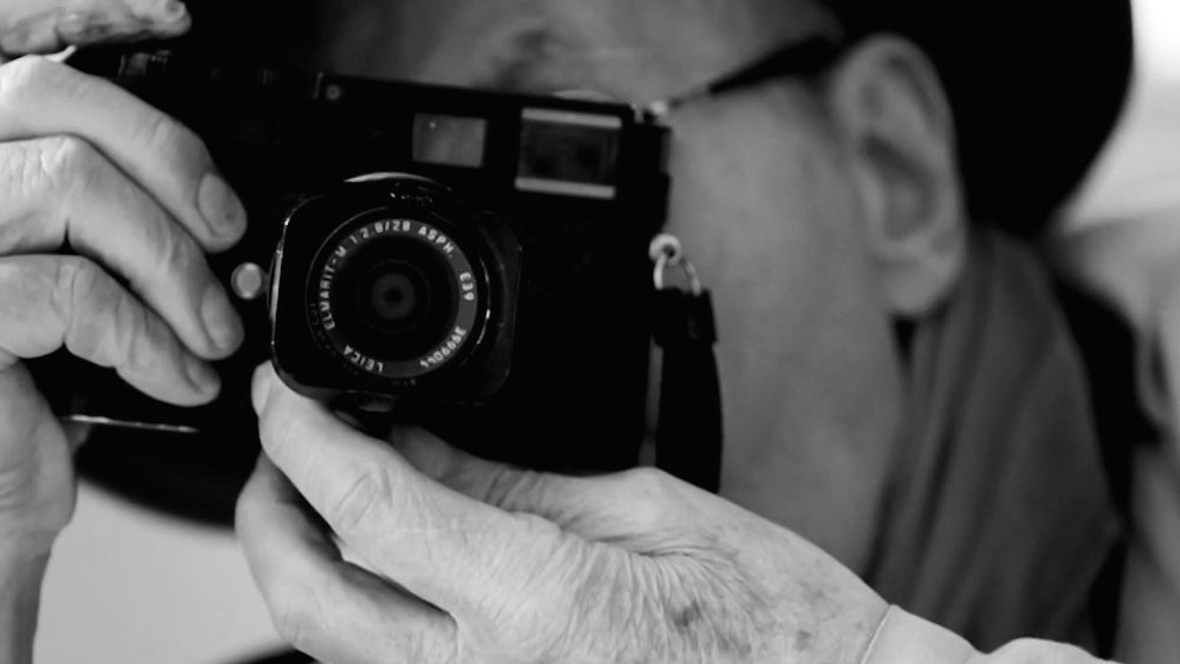 Black and white photo of a person holding a camera up to their face, focusing through the viewfinder, wearing glasses and a hat.