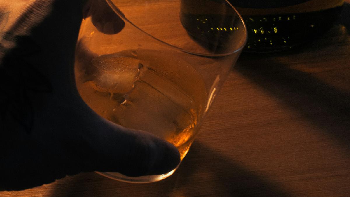 Photo of a hand holding a glass of whisky with ice next to a bottle on a wooden surface, dimly lit setting.