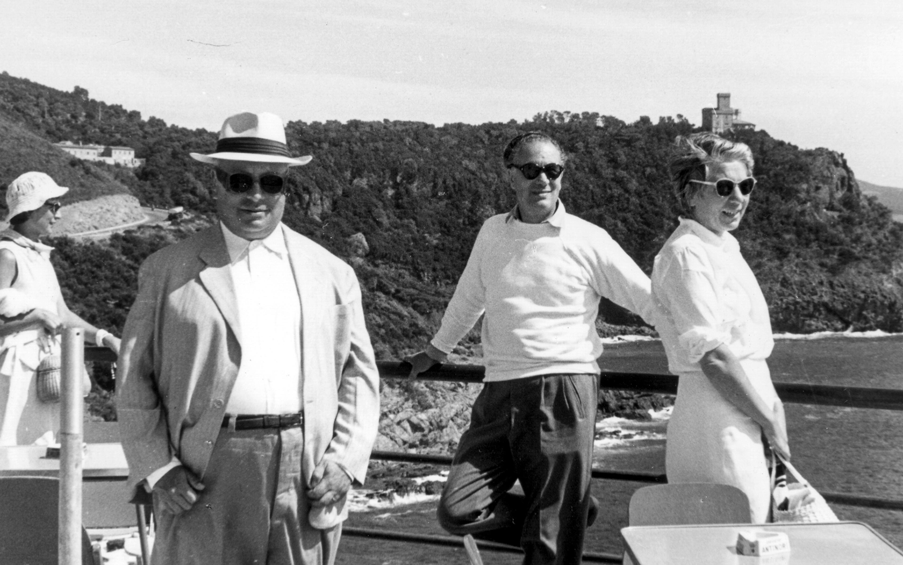 Black and white photo of four people in sunglasses standing on a terrace overlooking the sea with a hilly coastline in the background.