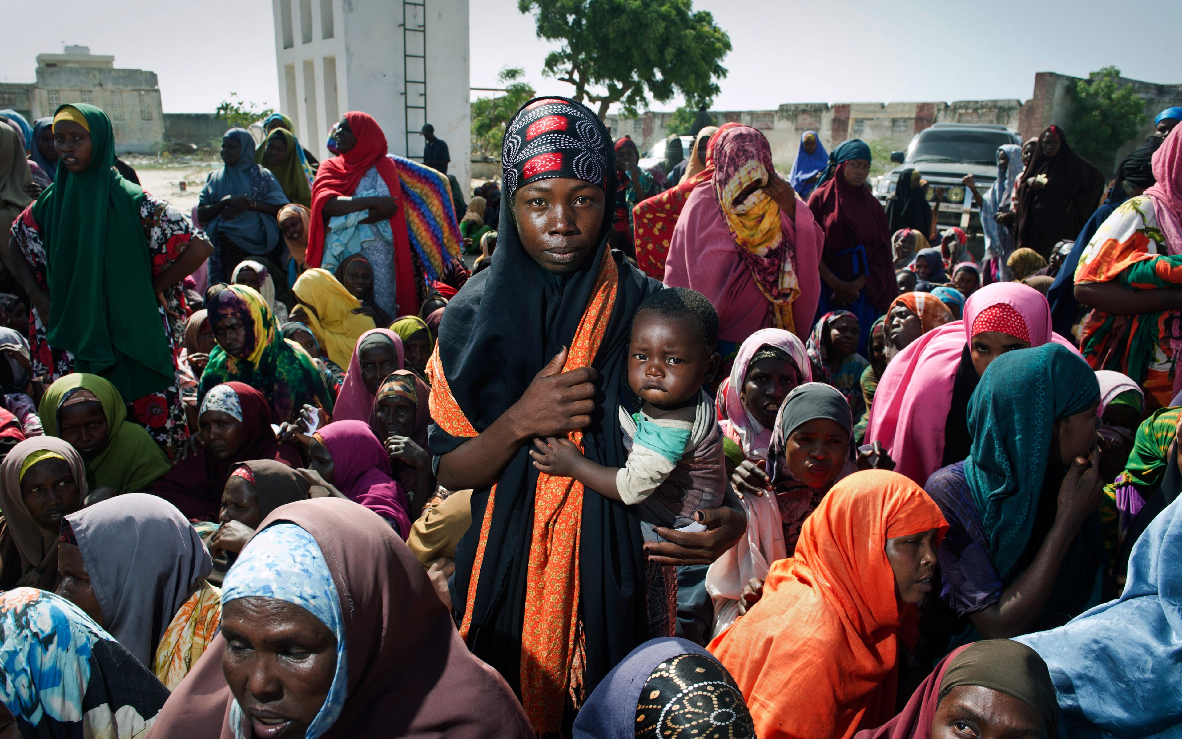 A woman holding a child in a crowded group, all wearing colourful clothing, outdoors with buildings and trees in the background.