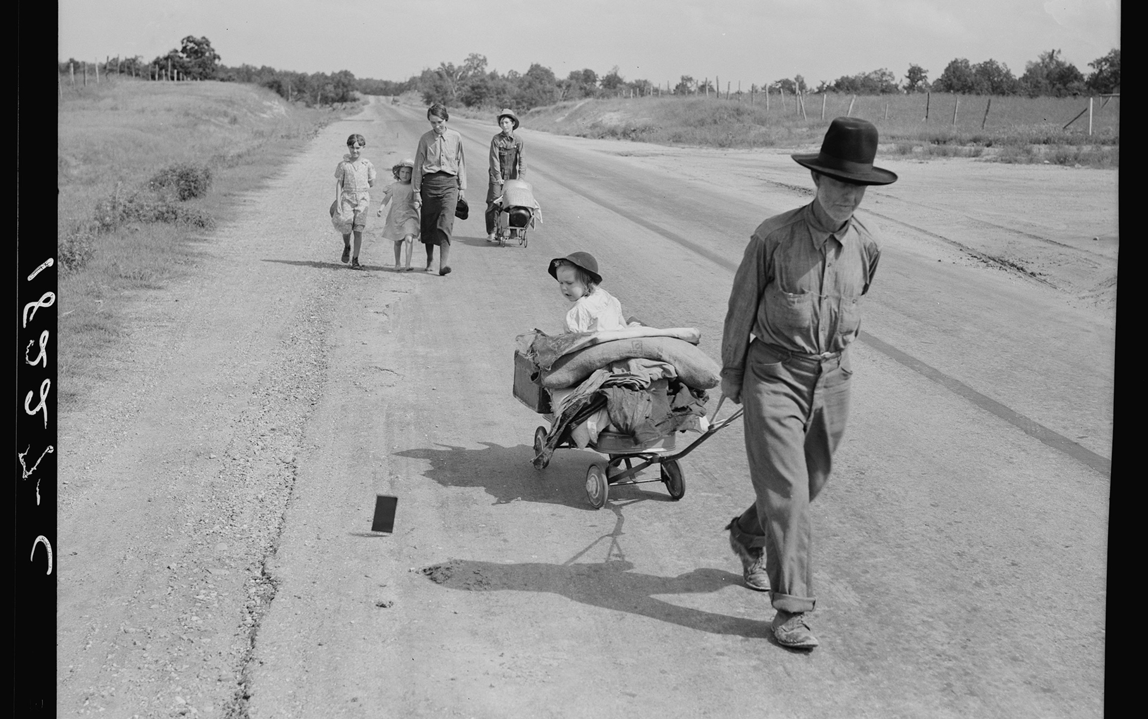 An old photograph of a man pulling a small cart with a child and belongings, followed by a woman and three children; one child is pushing a stroller.