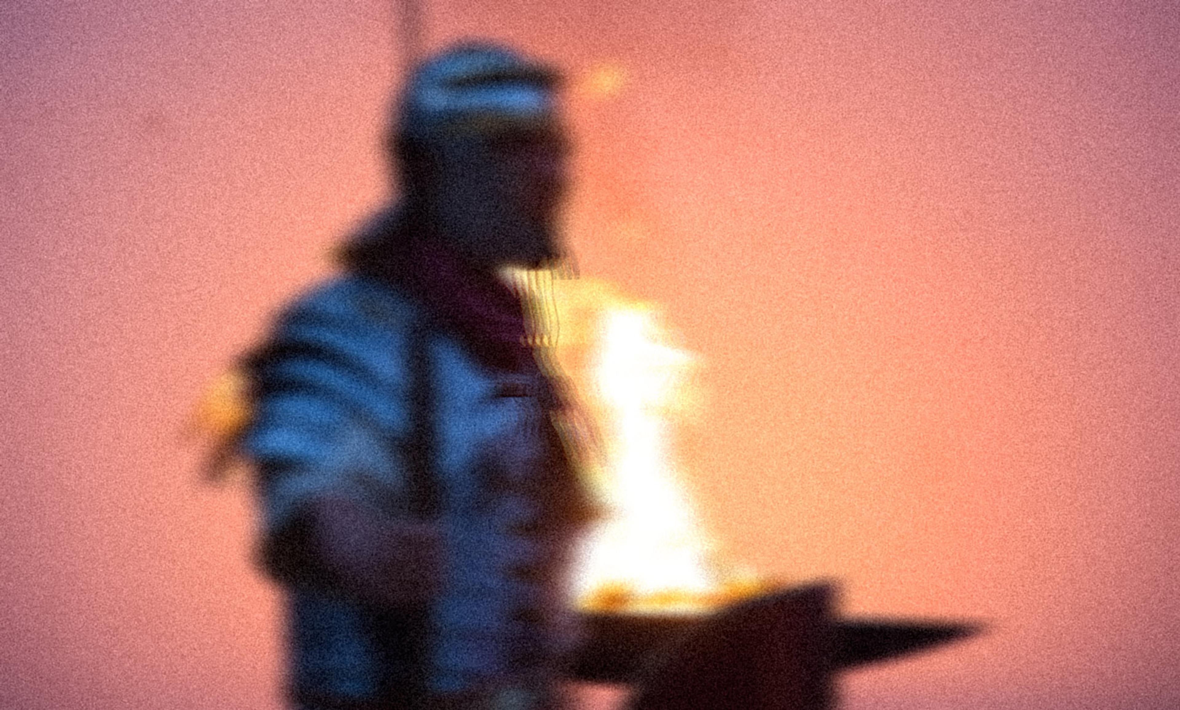 Blurry photo of a person wearing a headband and holding a torch, with a bright flame against a soft orange background.