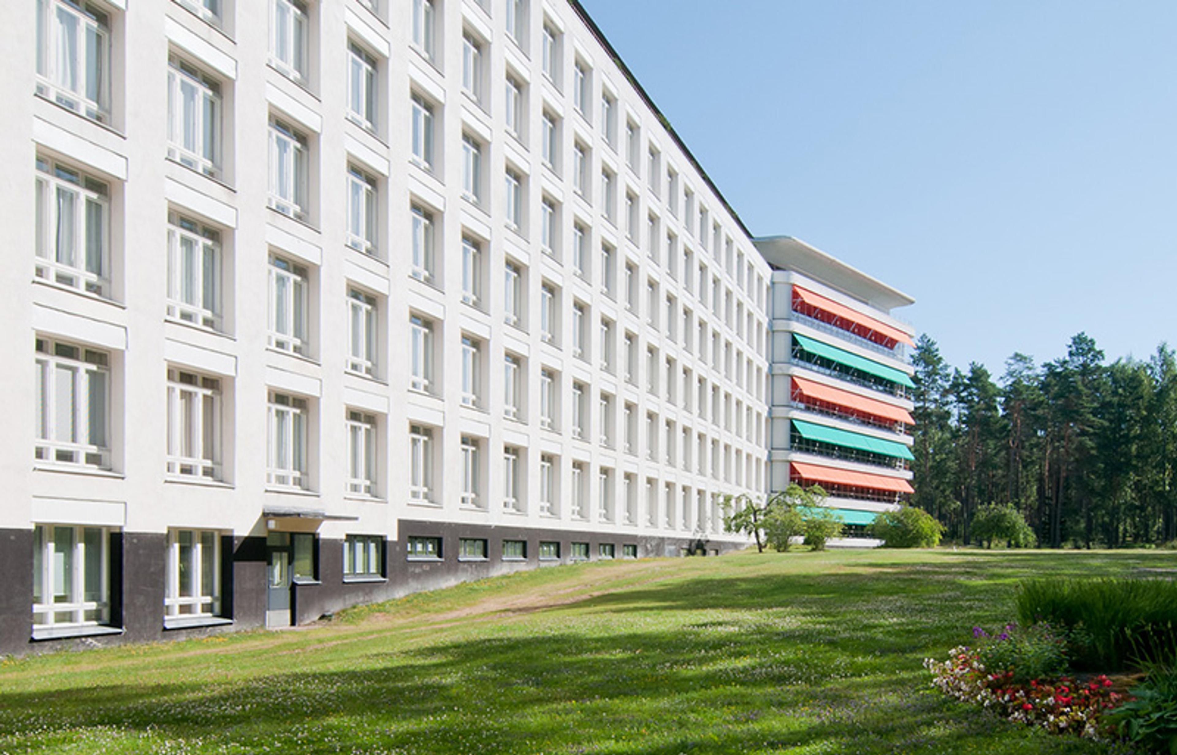 A white multi-storey building with many windows, green lawn in foreground, colourful balconies on the side, trees in background