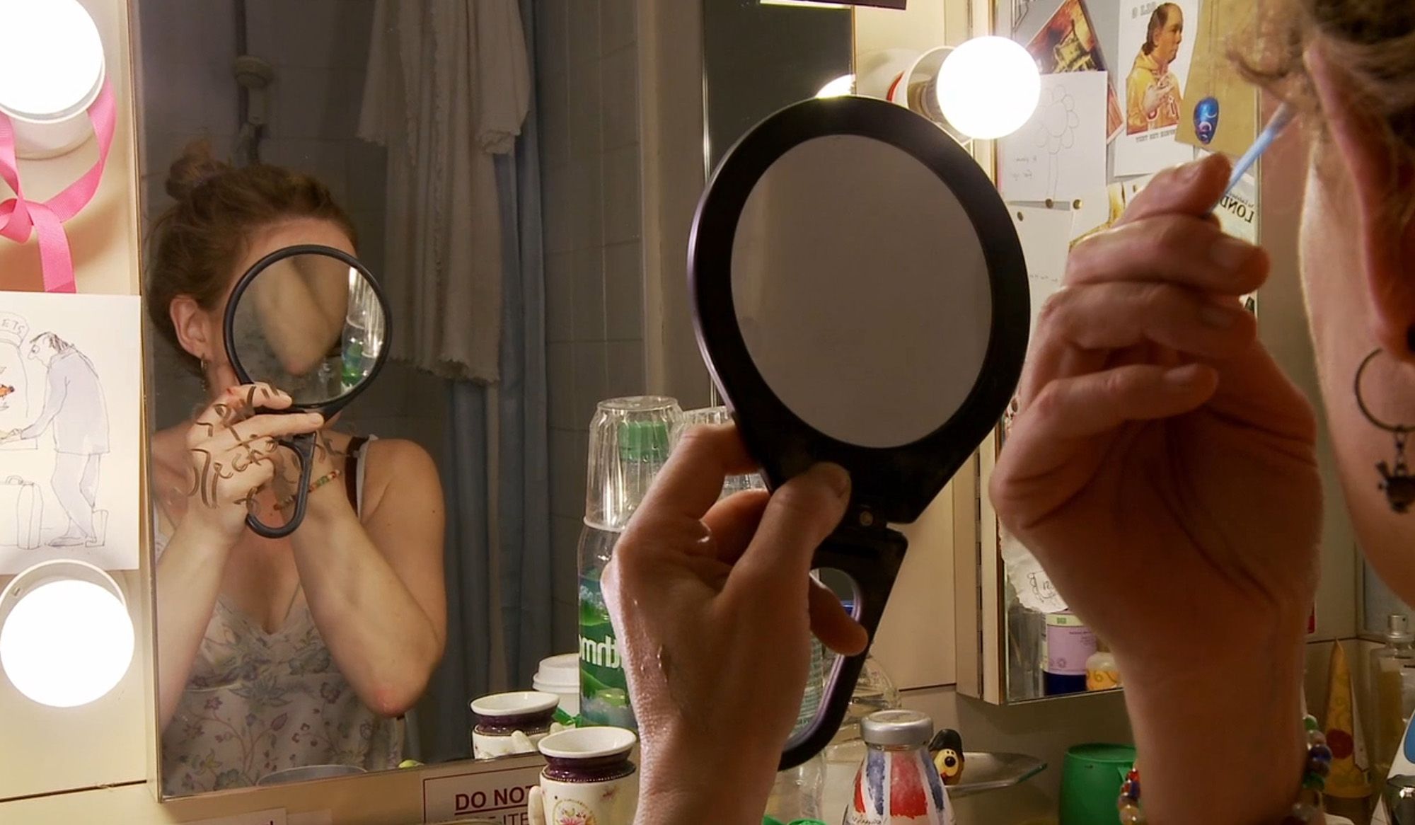 A woman holding a round mirror to her face while applying makeup in front of a dressing room mirror with lights and clutter.