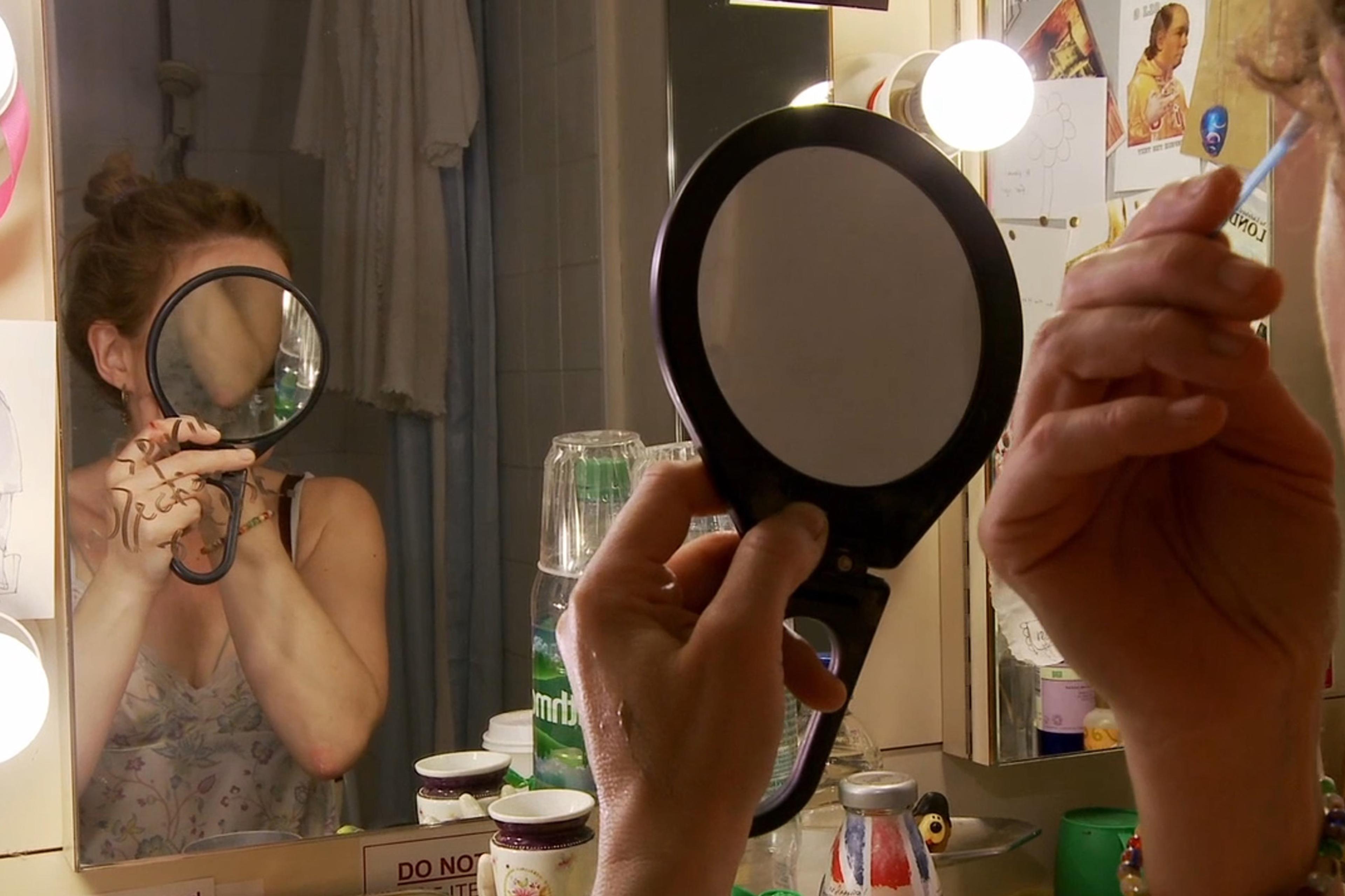 A woman holding a round mirror to her face while applying makeup in front of a dressing room mirror with lights and clutter.