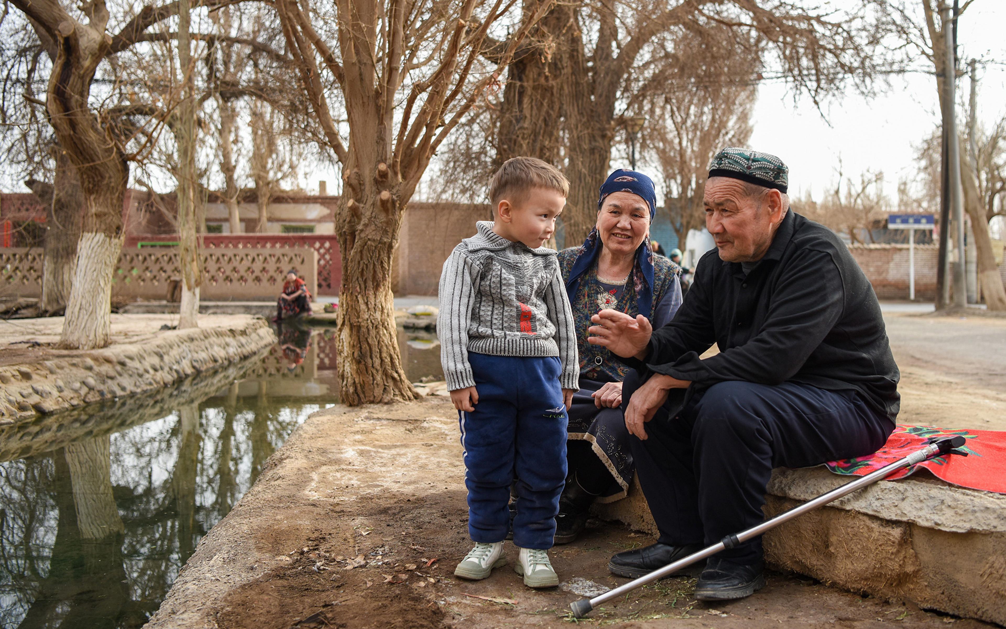 An elderly couple sit beside a child standing next to a canal with bare trees behind them. The man talks to the child and has a walking stick beside him.
