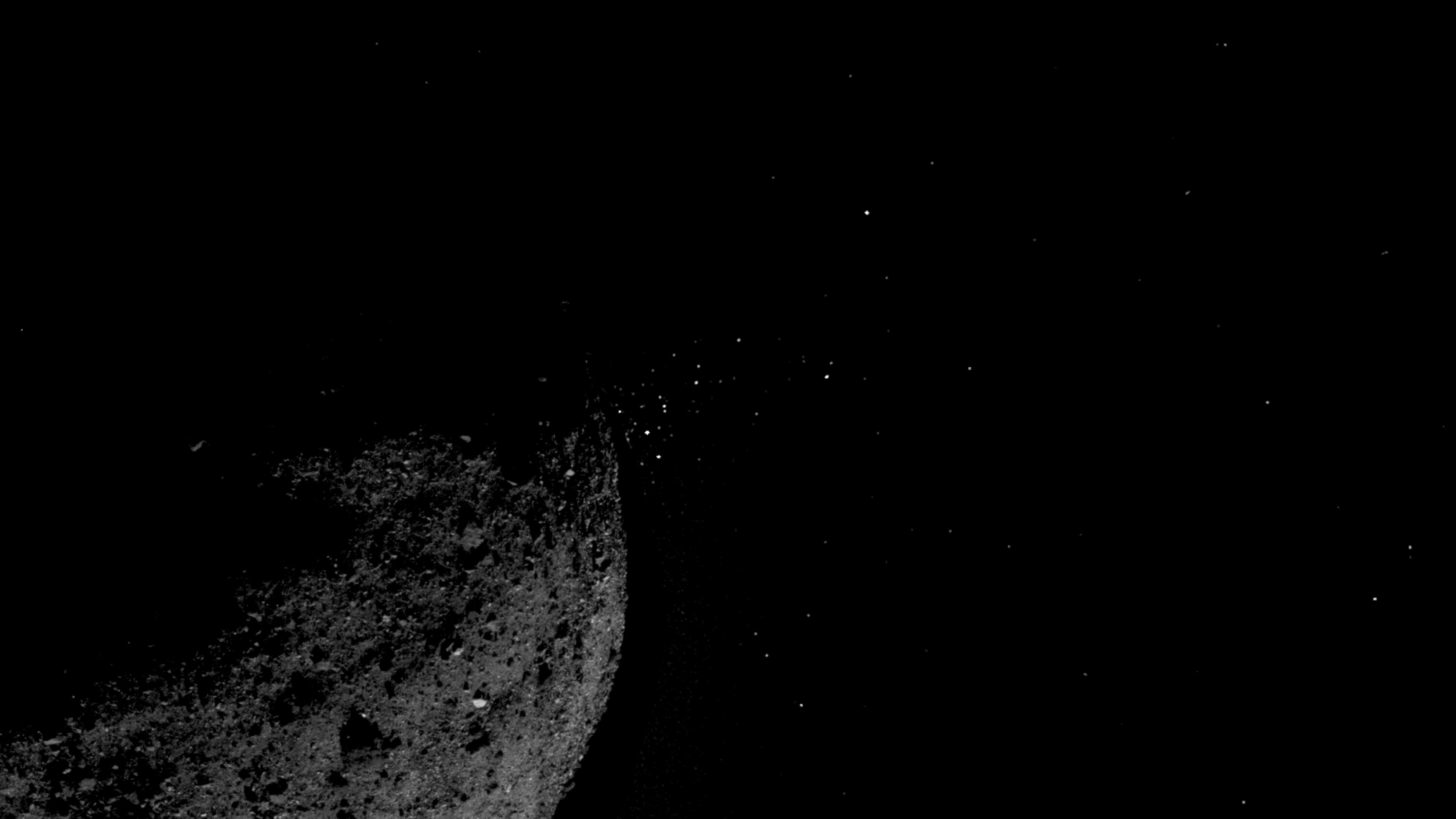 Photo of a dark rocky asteroid with a starry sky background and small particles visible near its surface.