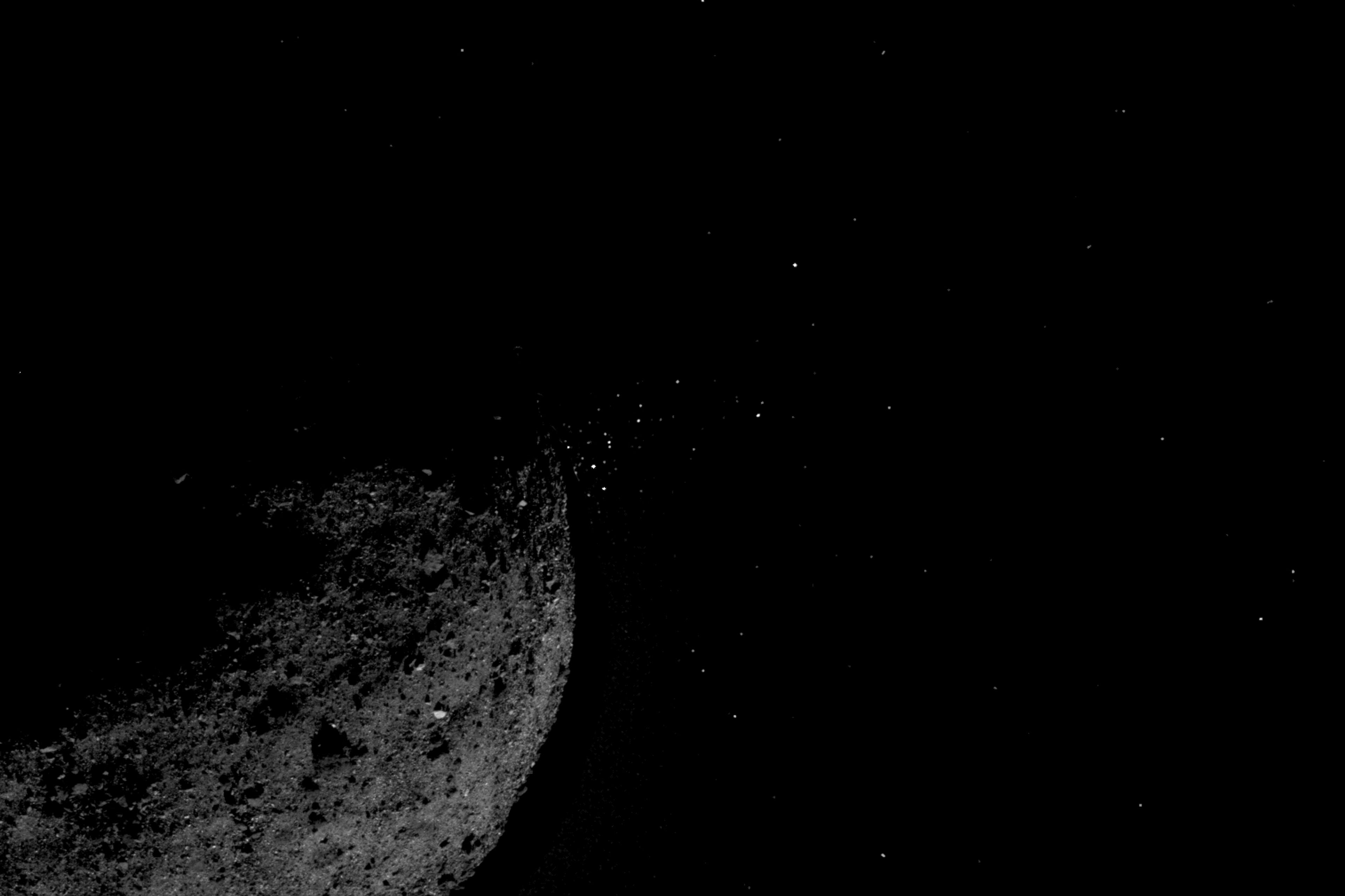 Photo of a dark rocky asteroid with a starry sky background and small particles visible near its surface.