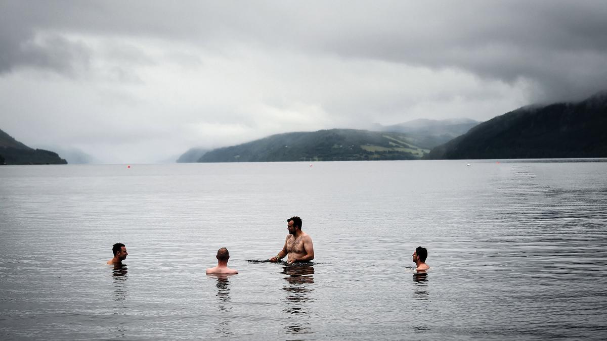 Four people swimming in a lake surrounded by foggy hills under a cloudy sky.