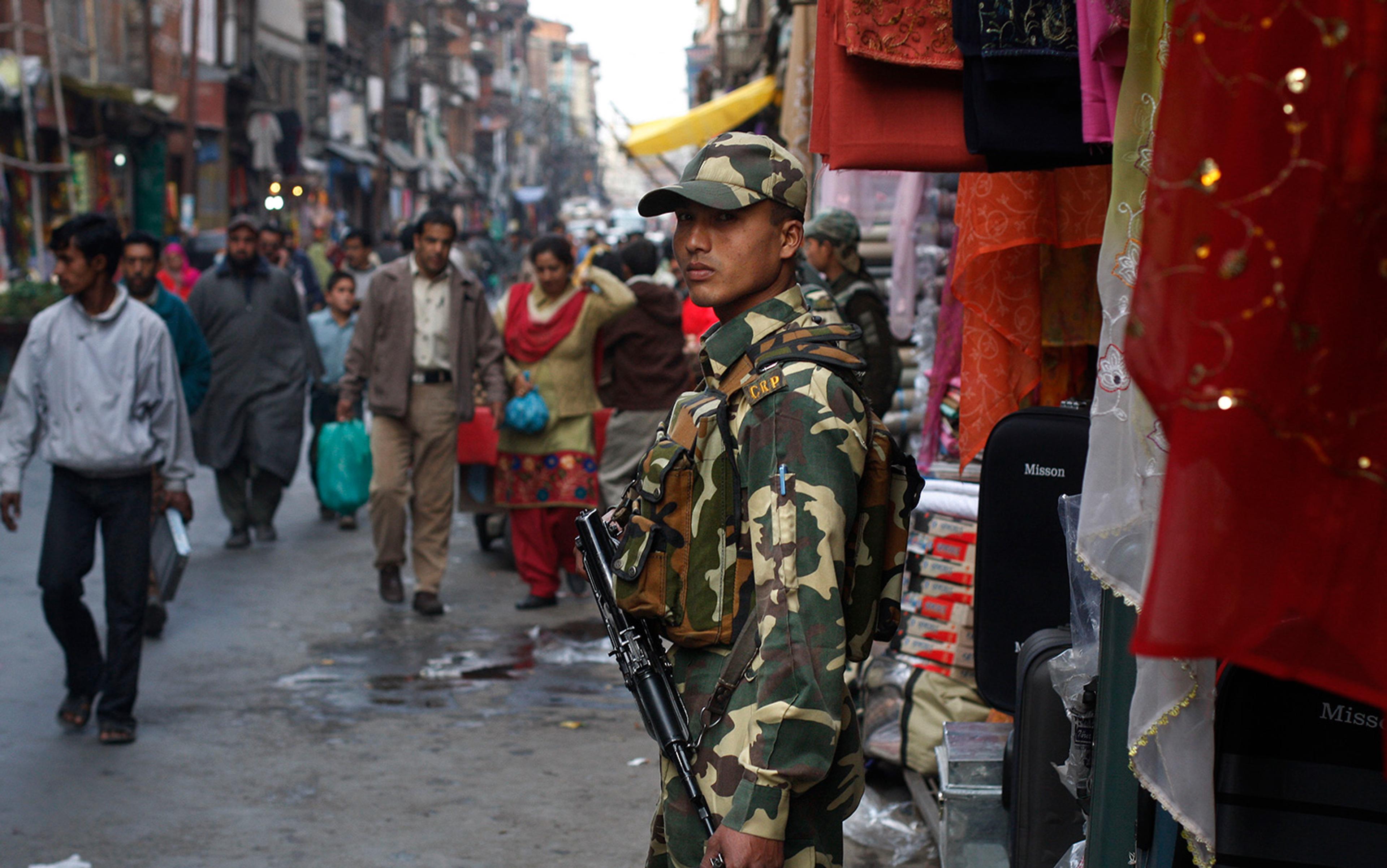 Photo of a soldier in camouflage standing on a busy market street surrounded by people and colourful fabrics.