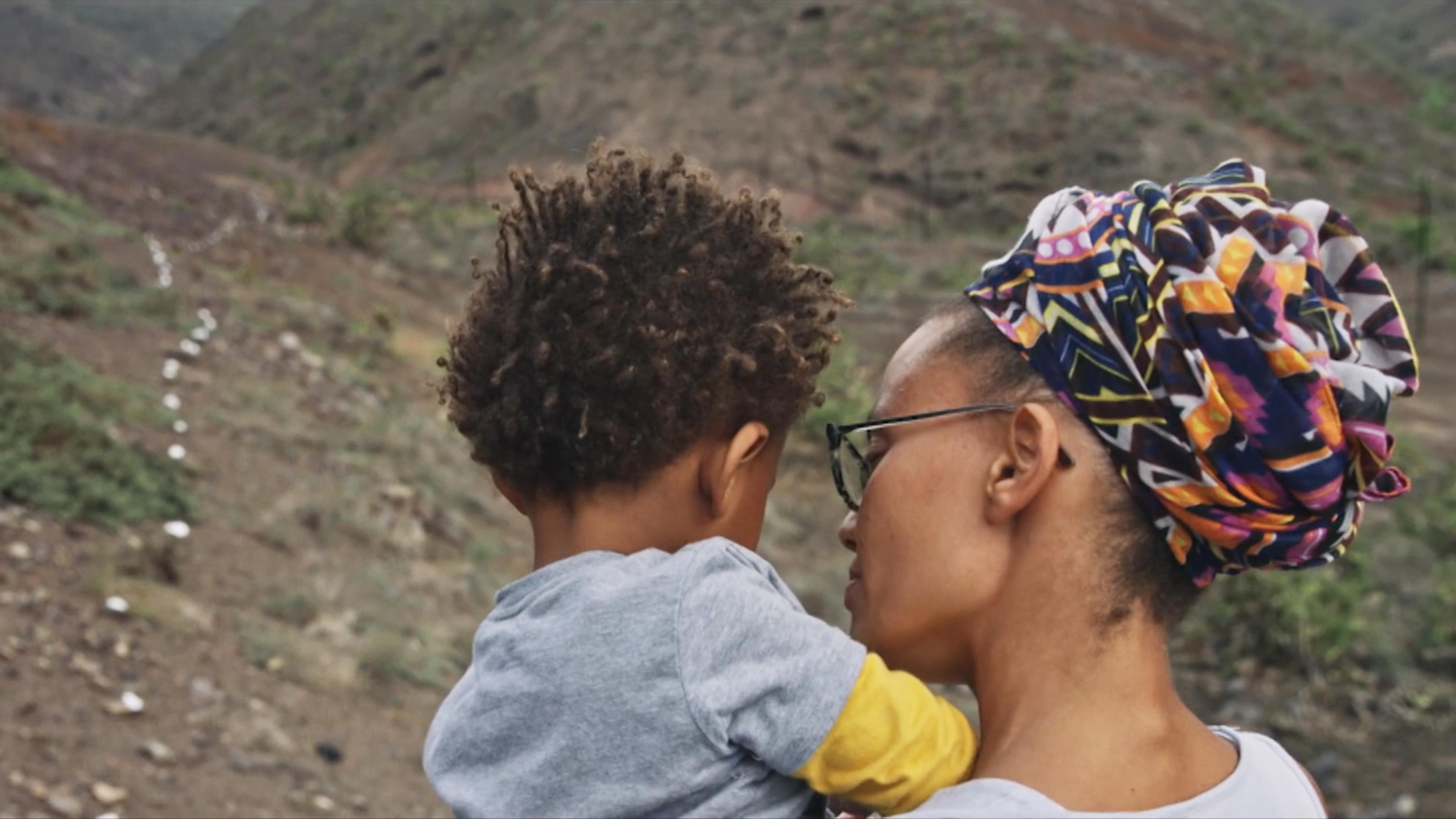 A woman with glasses and a colourful African headscarf is holding a child. They are outdoors in a rocky, hilly landscape. The path ahead appears scattered with small white stones.