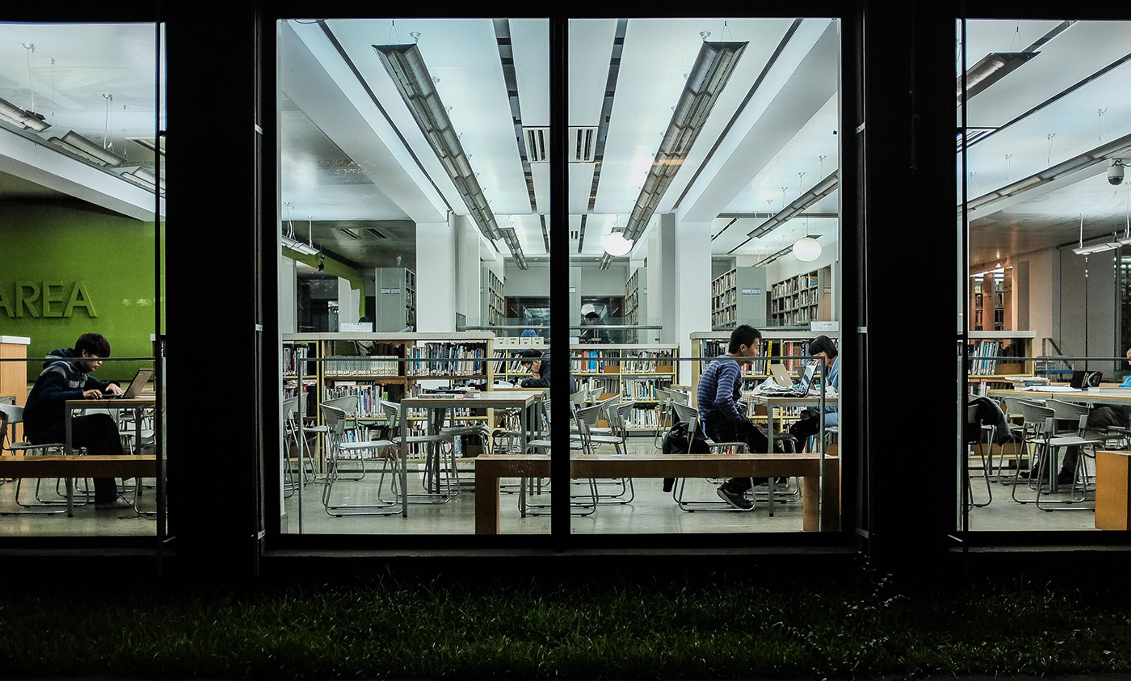 People studying with laptops in a modern library, viewed at night through large windows. Shelves of books are visible.