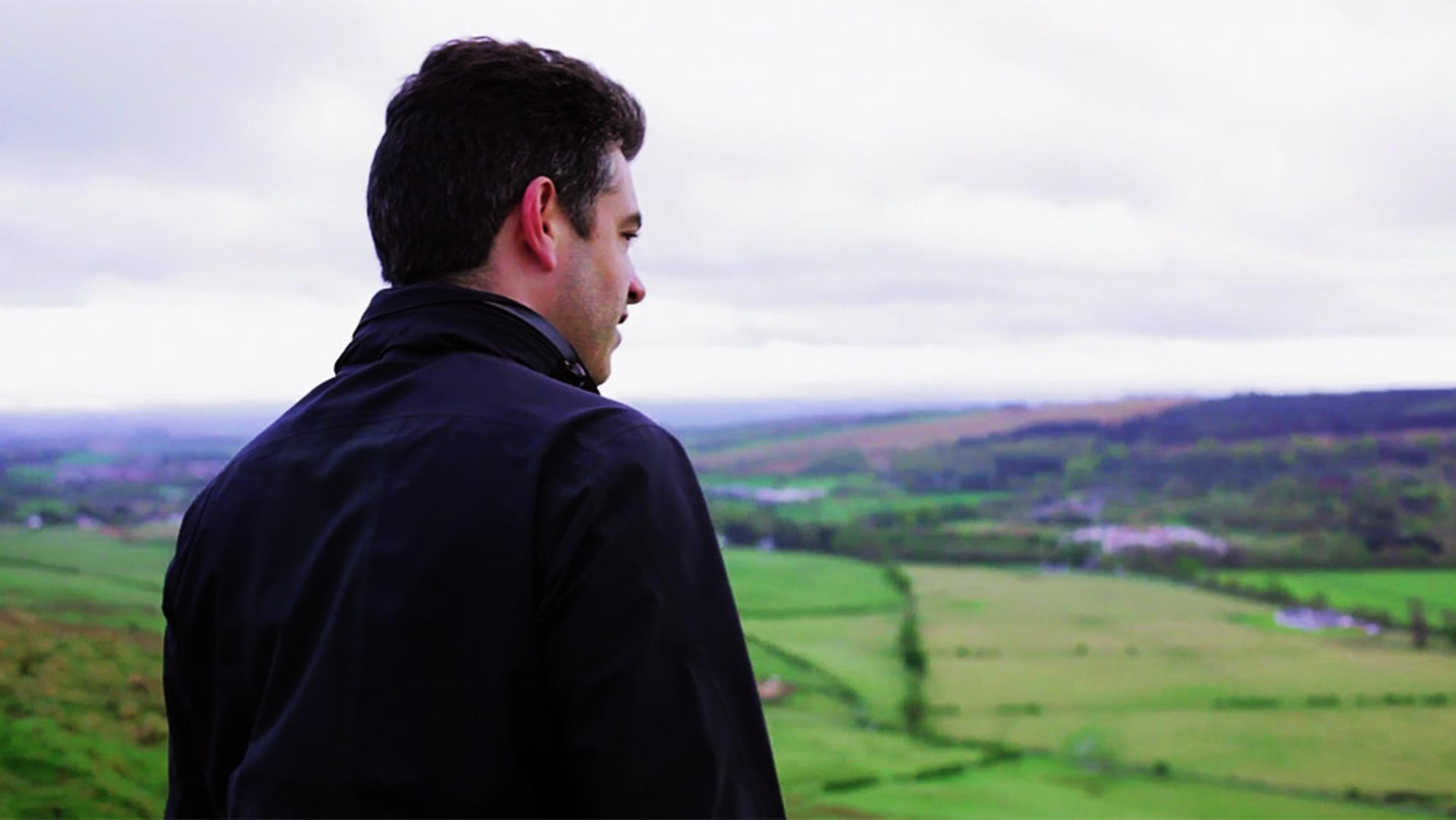 A man with dark hair in a black jacket looking over a vast green landscape under a cloudy sky.
