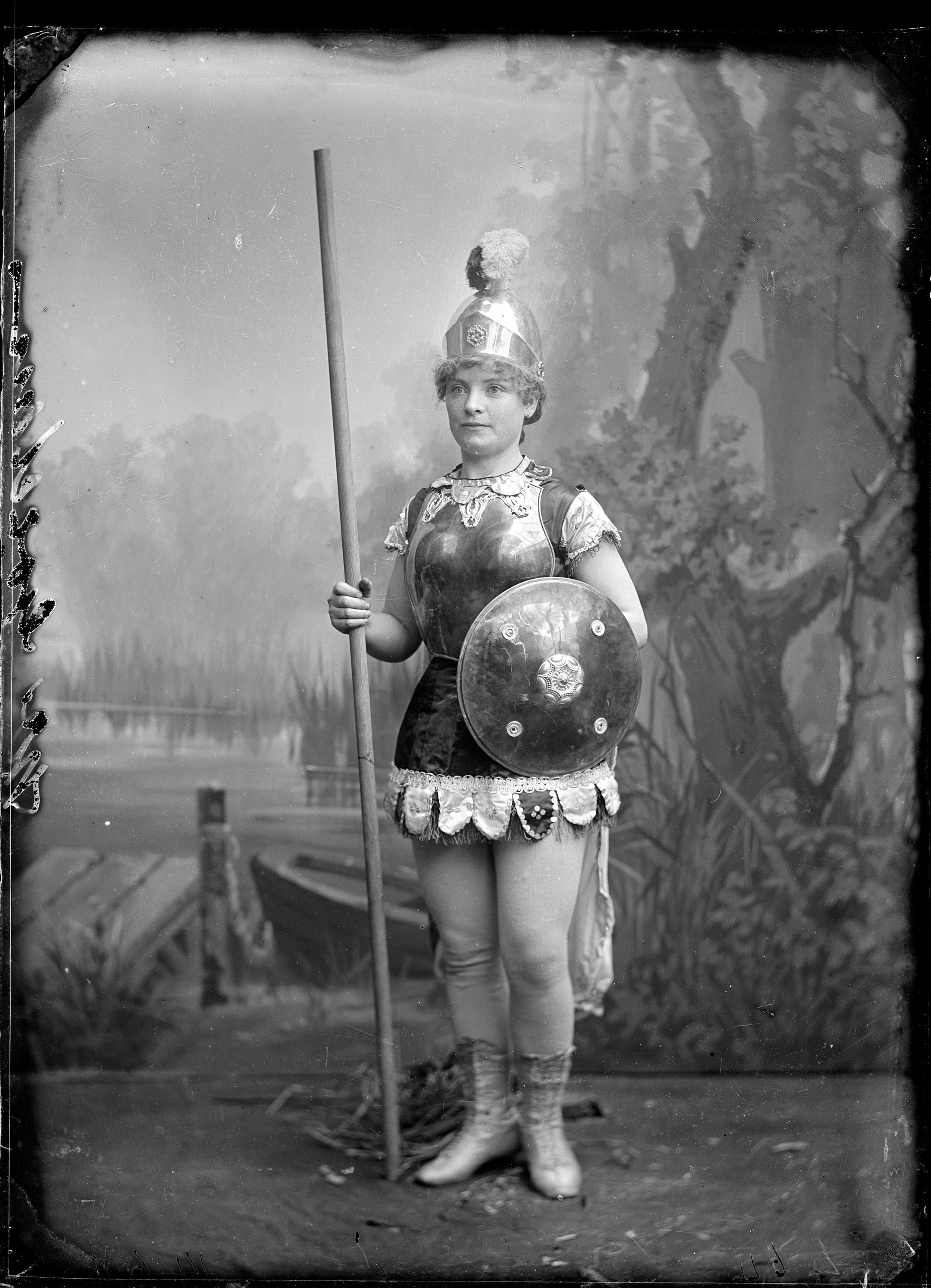 Black and white photo of a woman in a historic costume with a spear and shield posing in front of a painted forest backdrop.