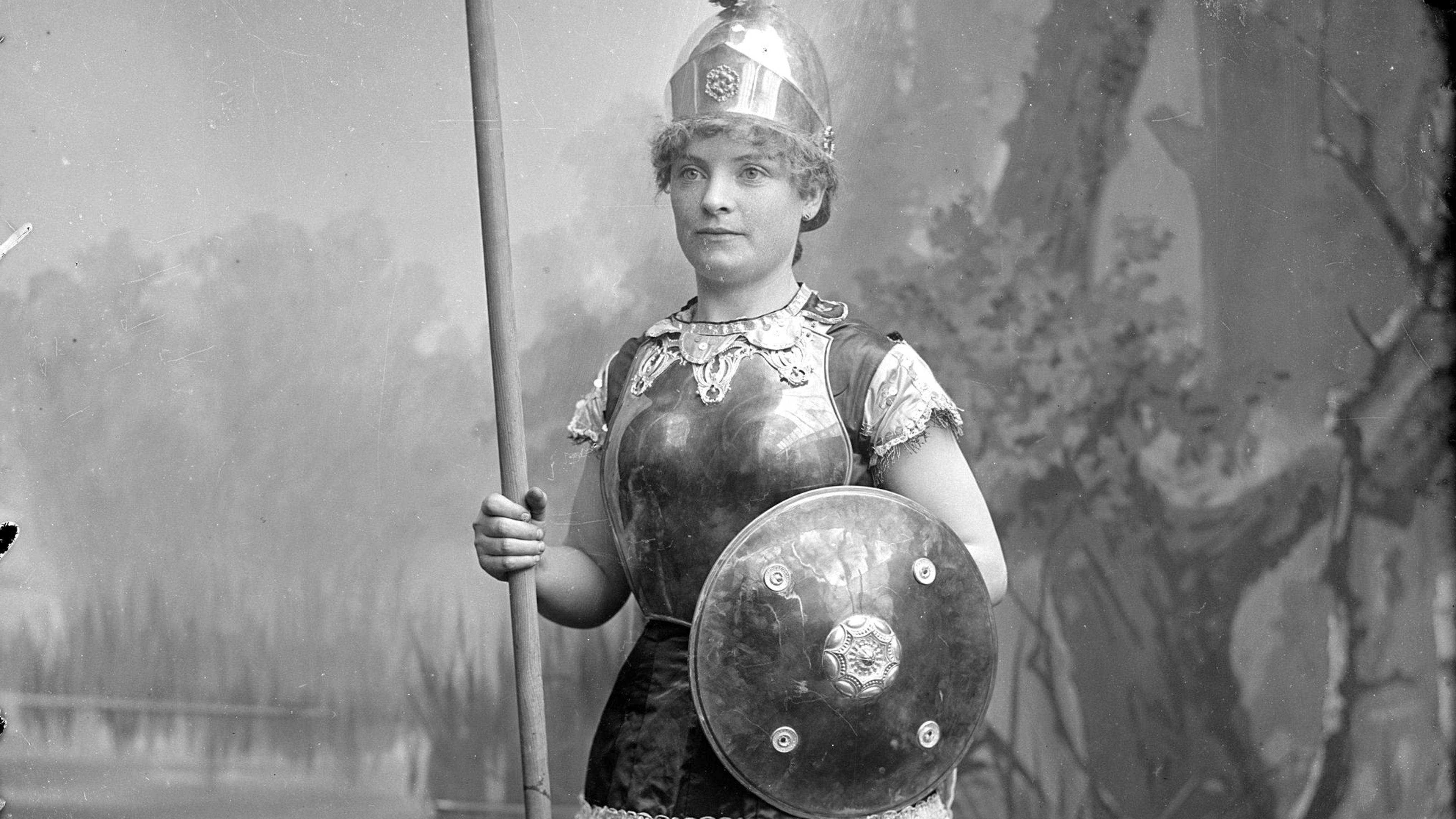 Black and white photo of a woman in a historic costume with a spear and shield posing in front of a painted forest backdrop.