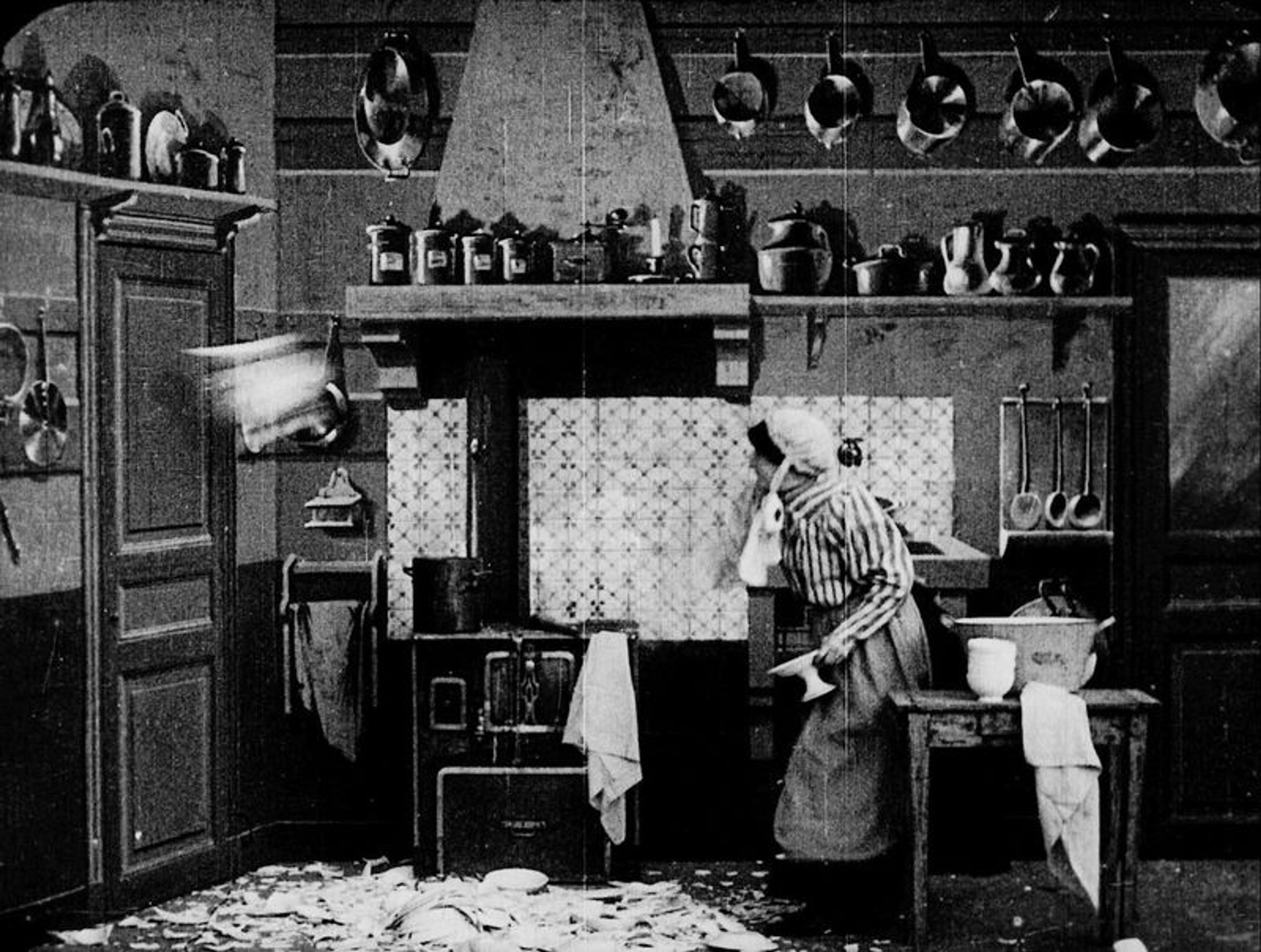 Black and white photo of a vintage kitchen with a woman cleaning up broken dishes; pots hang from the wall were knocked down.
