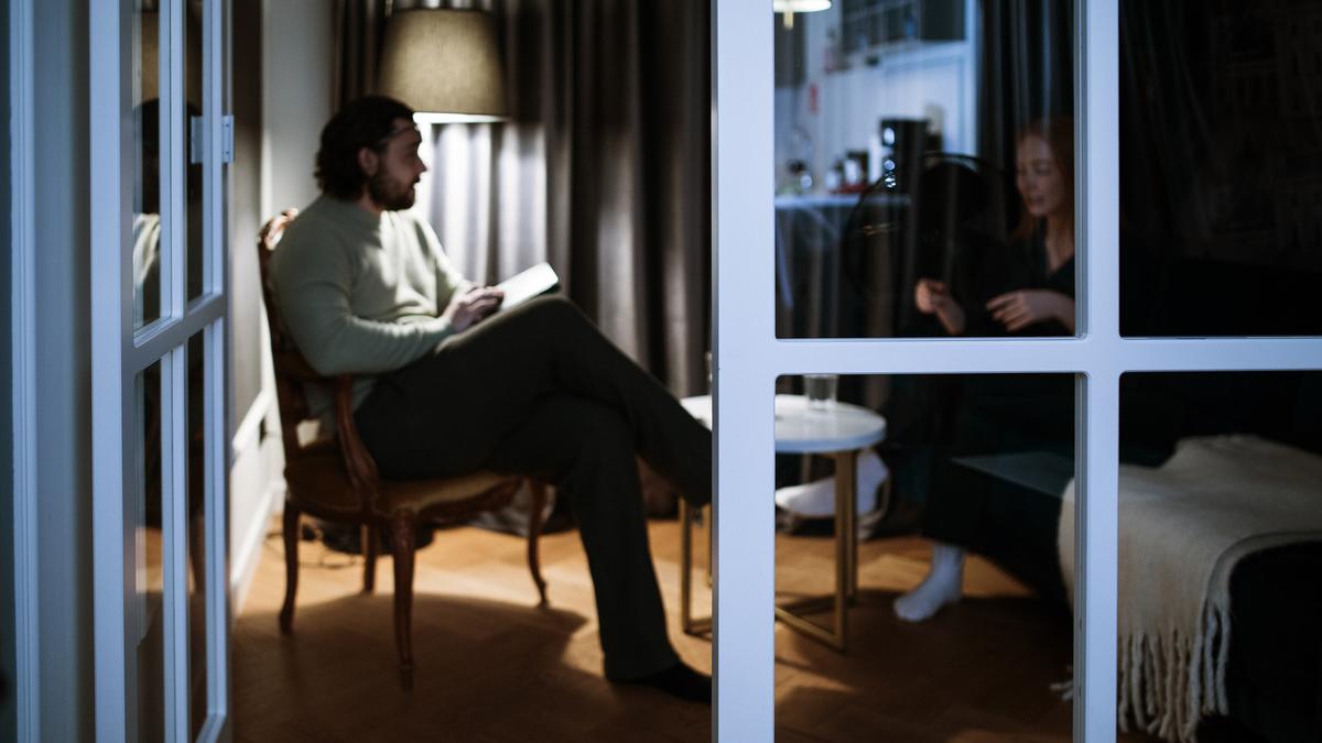 A man holding a notepad and a woman talking in a dimly lit room seen through glass doors.