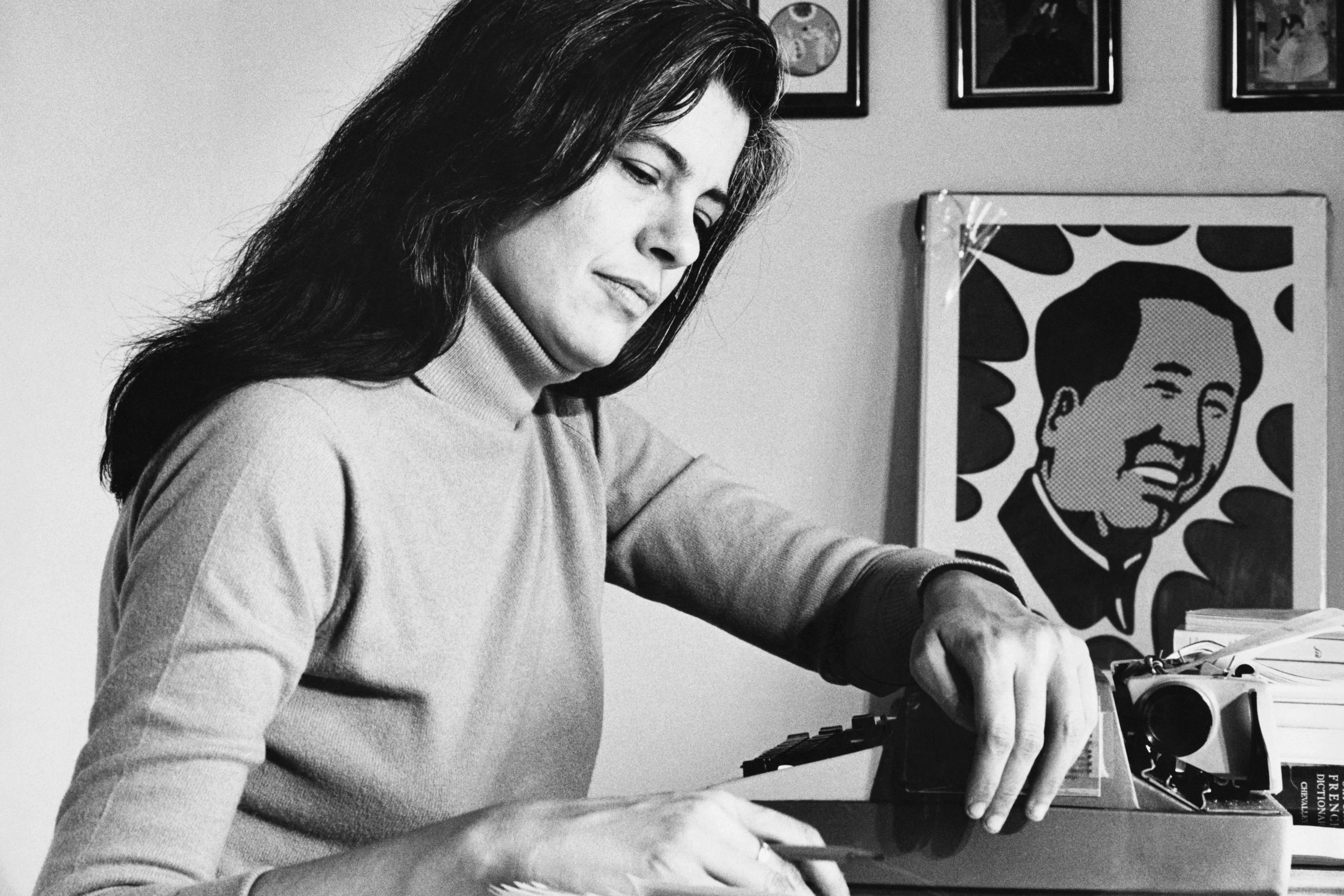 Black and white photo of a woman typing at a desk with a poster of Mao Zedong and framed art on the wall.