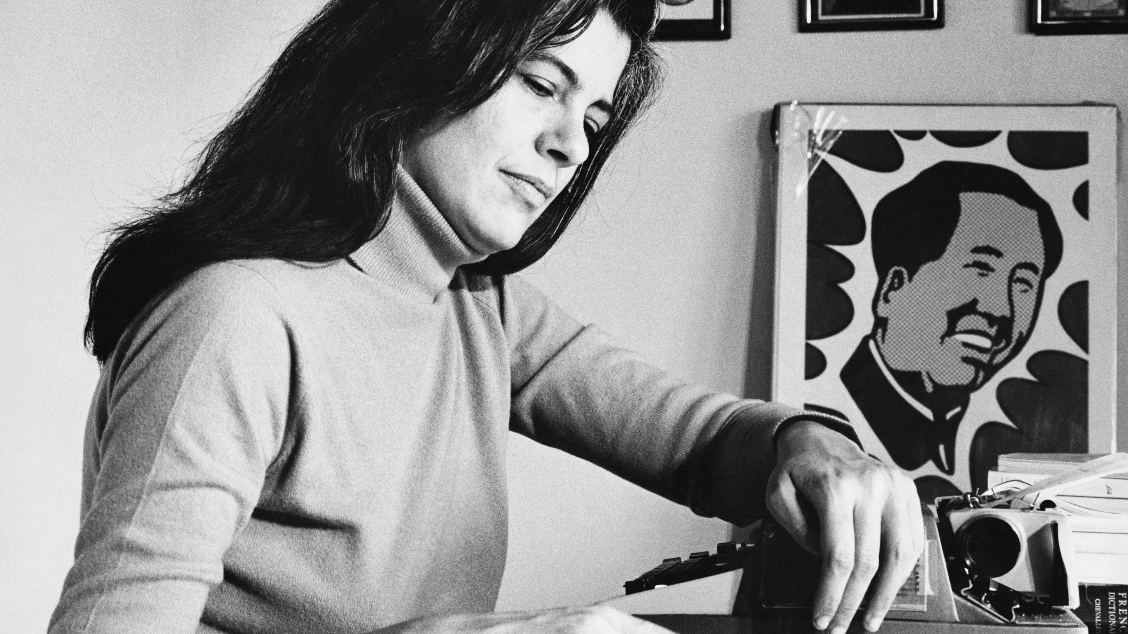 Black and white photo of a woman typing at a desk with a poster of Mao Zedong and framed art on the wall.