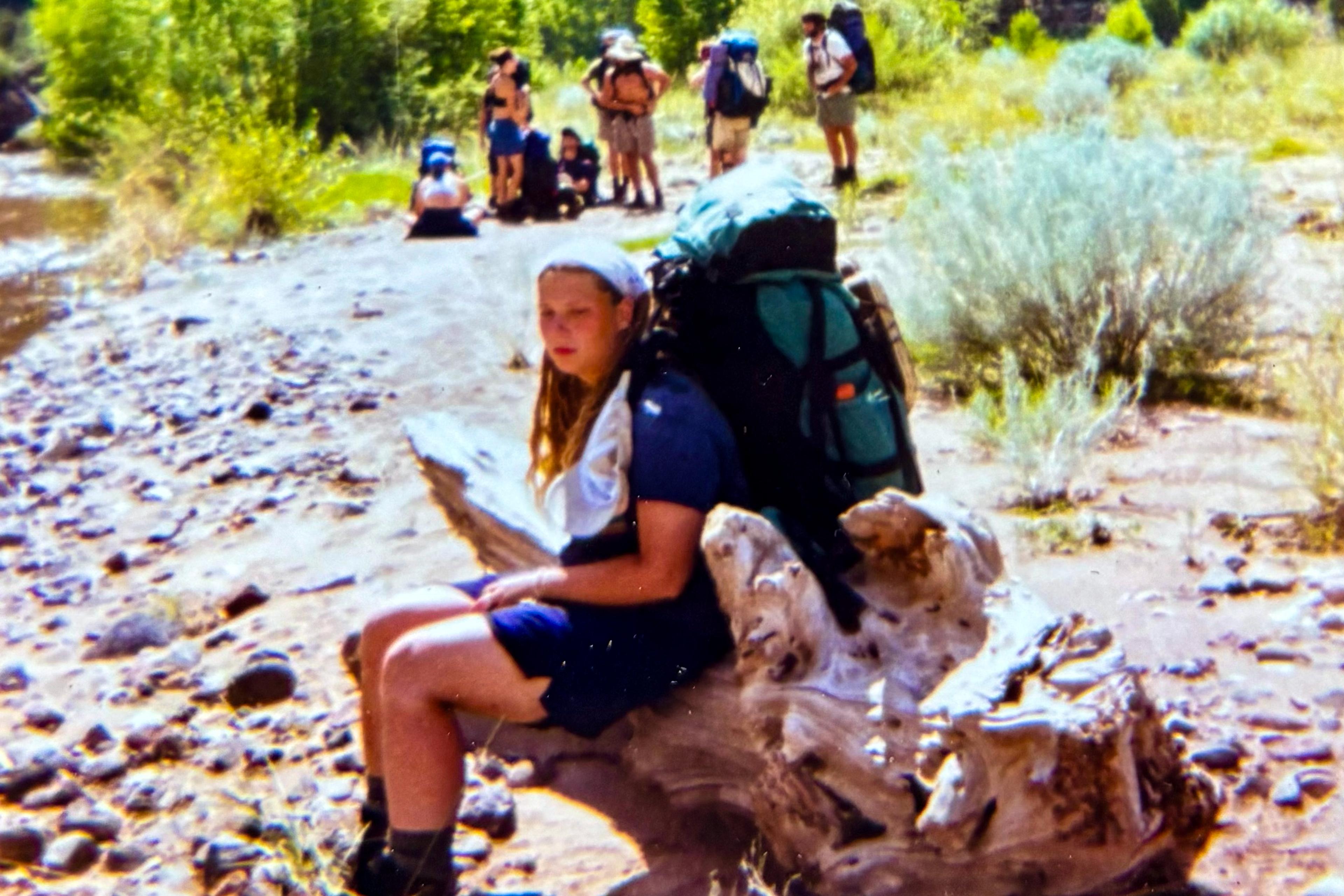 A person with a backpack resting on a log by a rocky riverbank with a group of people in the background.
