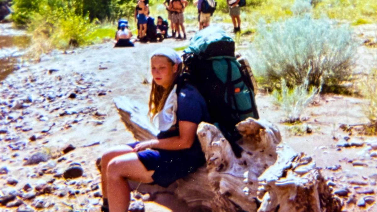 A person with a backpack resting on a log by a rocky riverbank with a group of people in the background.