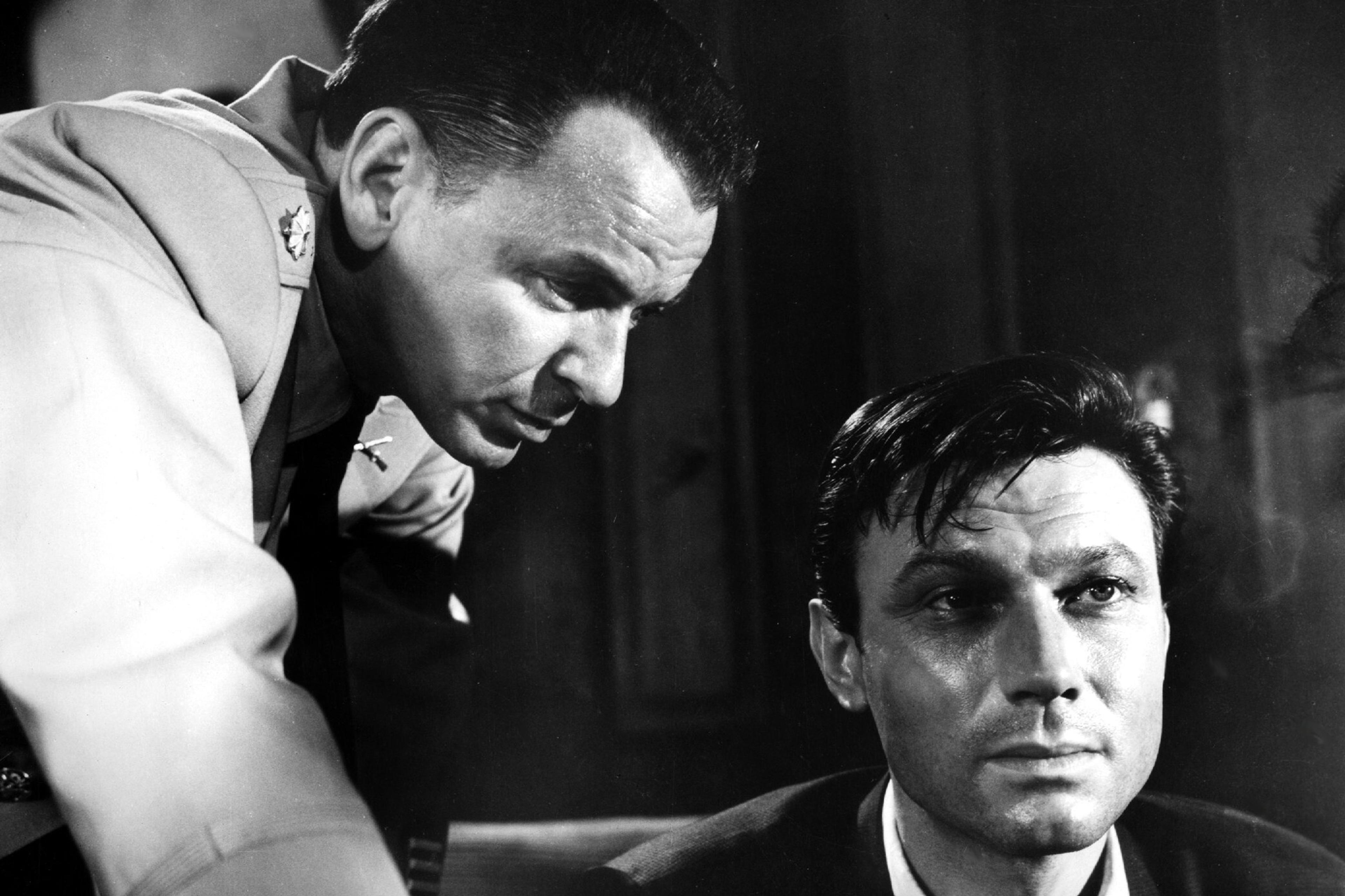 Black and white photo of two men, one in military uniform leaning over the other, who sits looking serious.