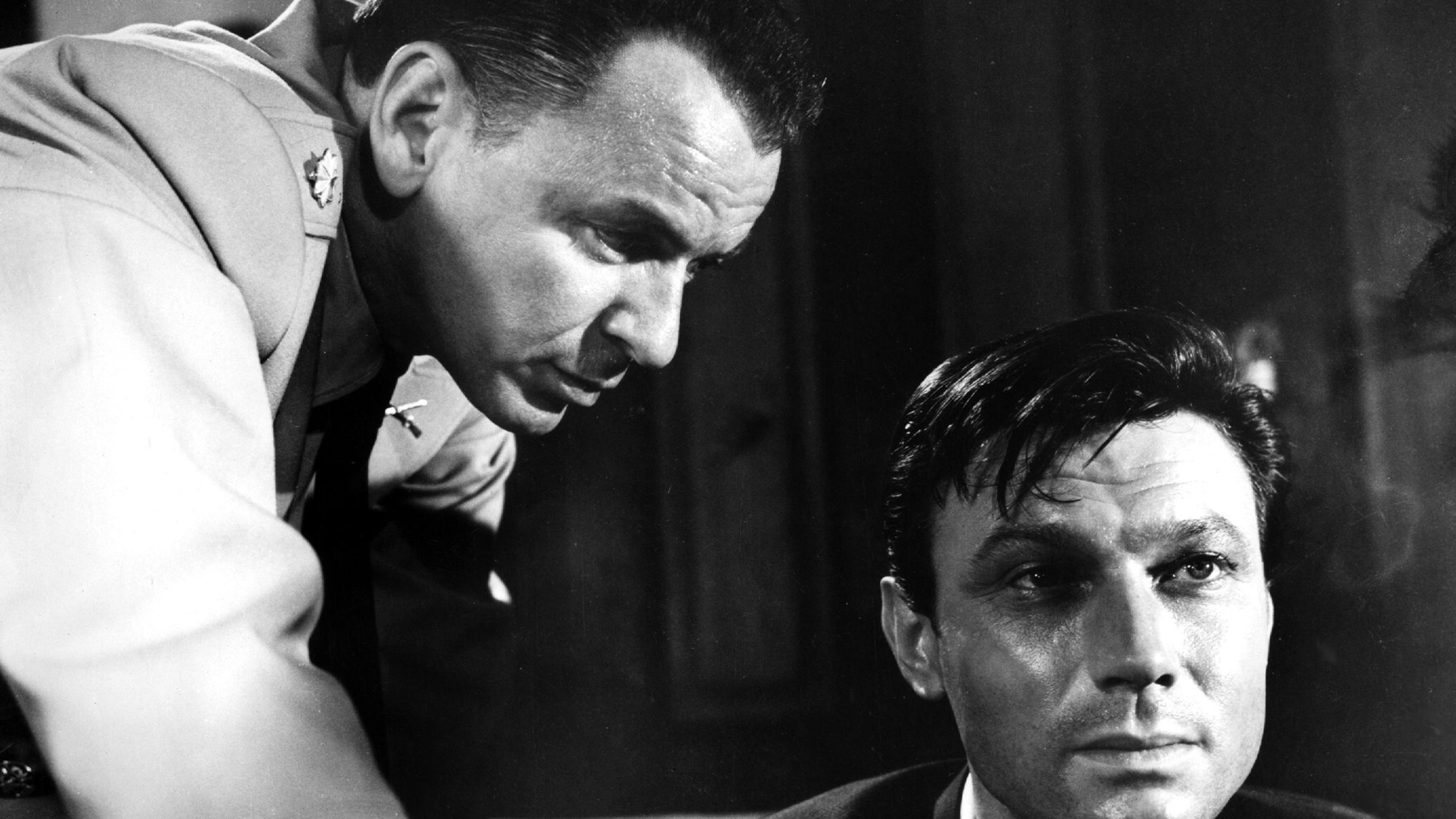 Black and white photo of two men, one in military uniform leaning over the other, who sits looking serious.