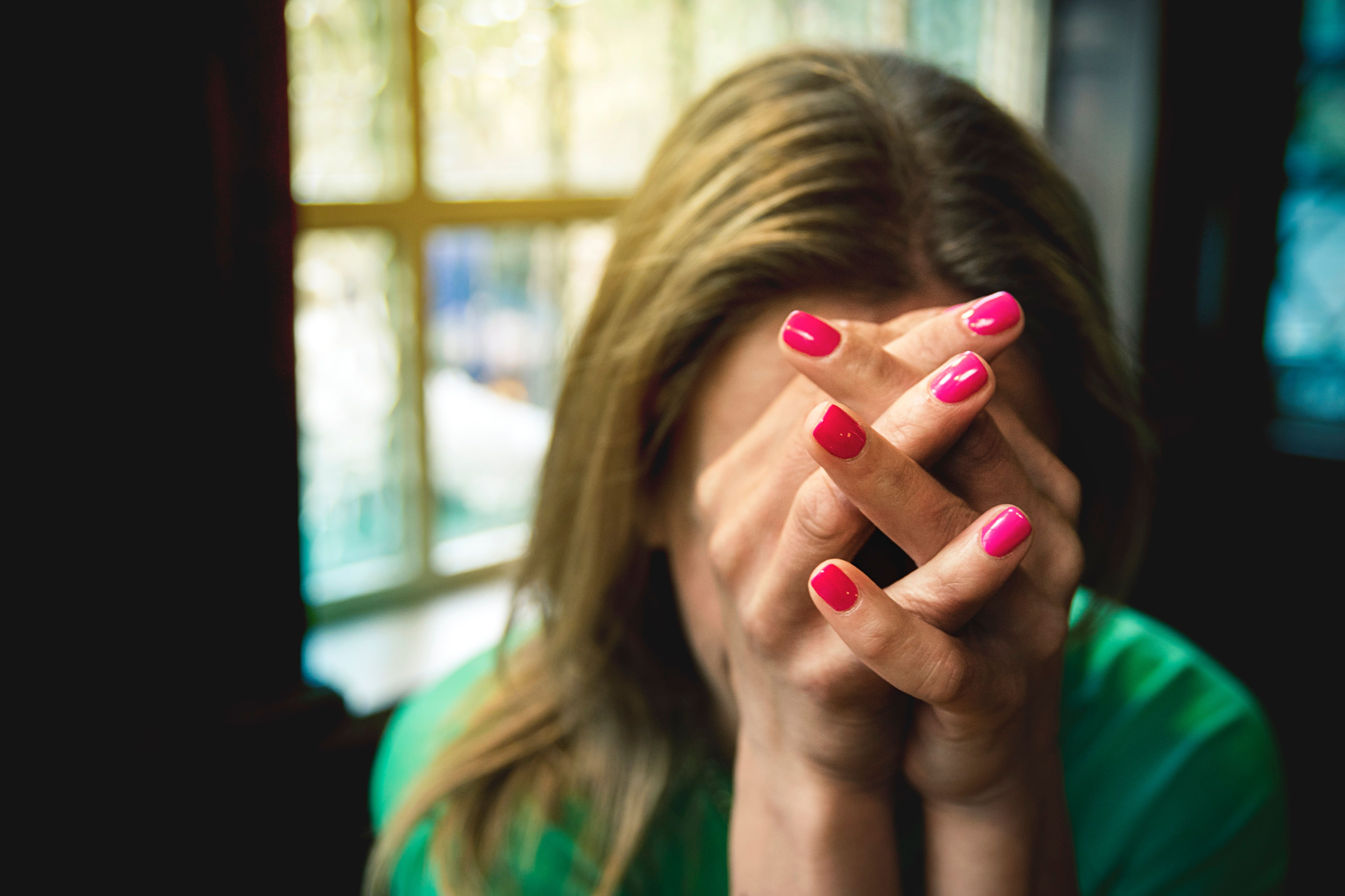 Photo of a person with pink nails covering their face with clasped hands near a window.