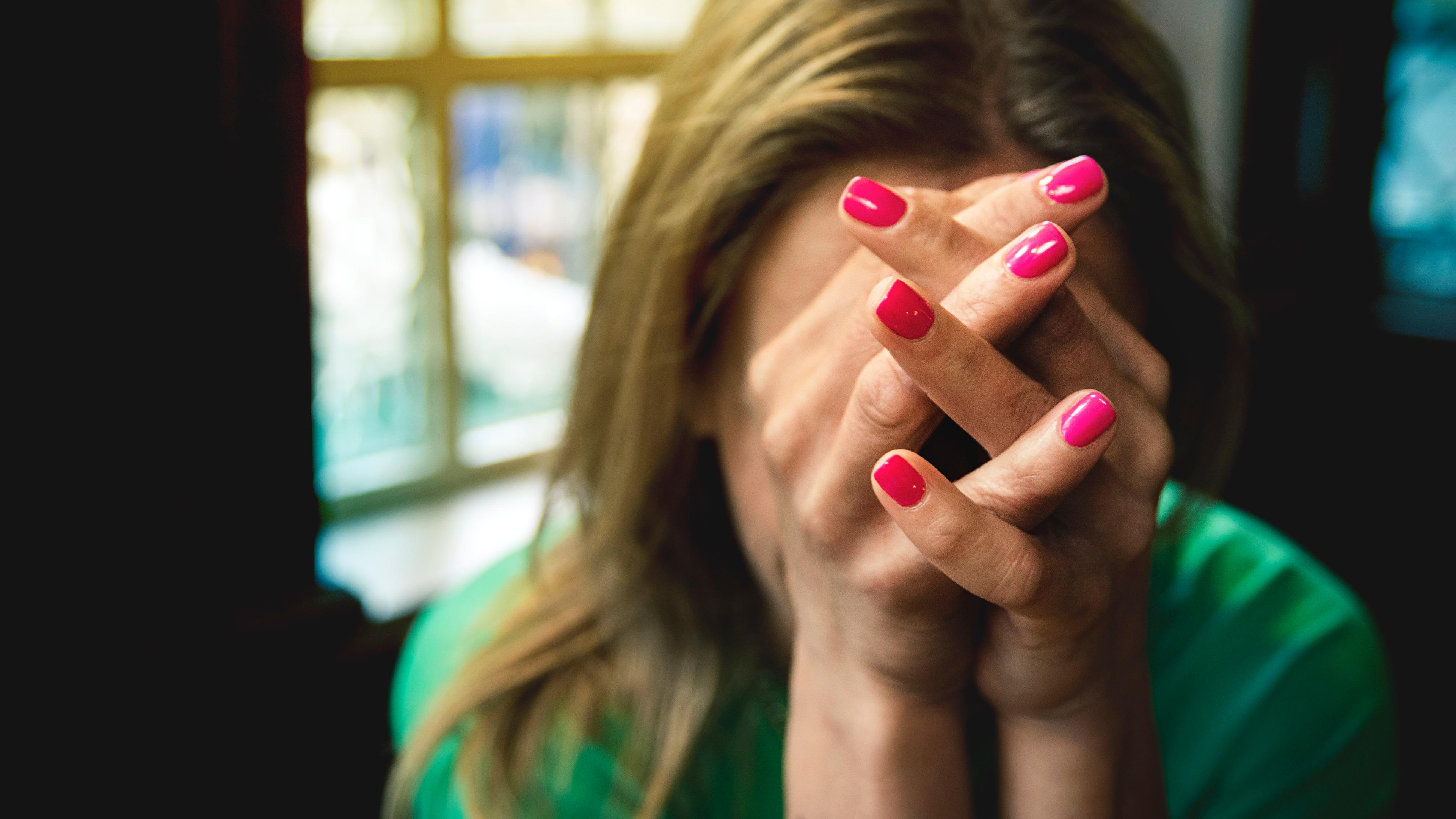 Photo of a person with pink nails covering their face with clasped hands near a window.