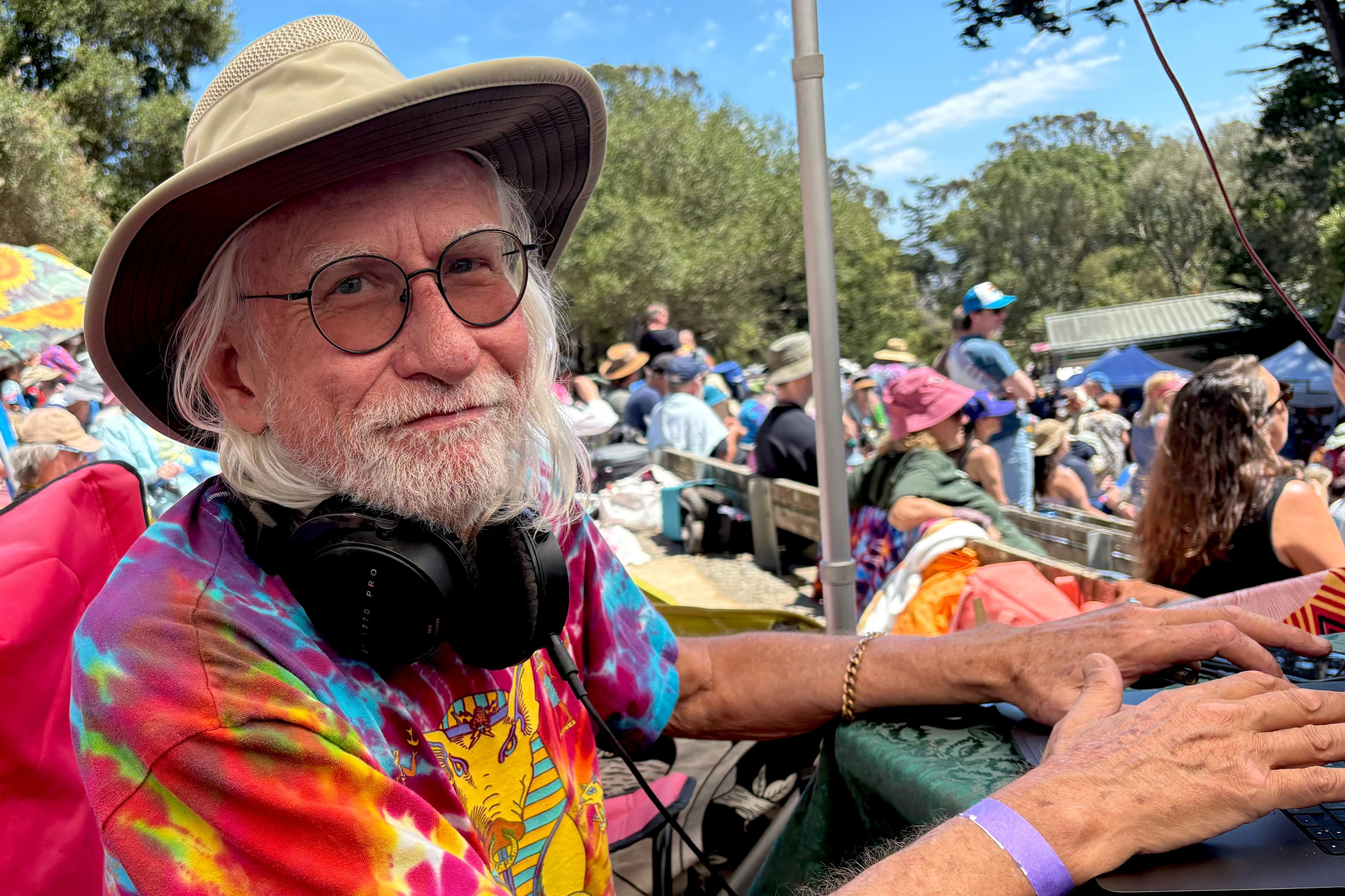 An older man in a hat and glasses with a colourful tie-dye shirt at an outdoor event, headphones around his neck.