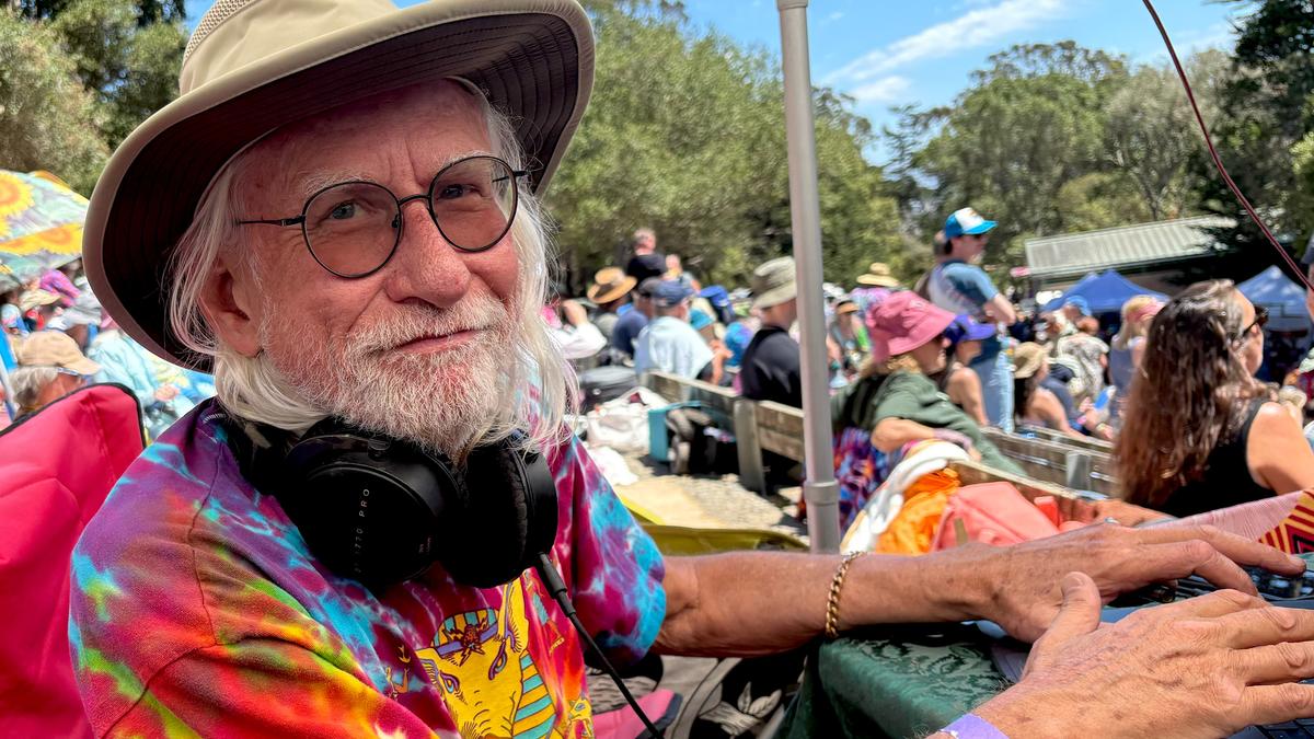 An older man in a hat and glasses with a colourful tie-dye shirt at an outdoor event, headphones around his neck.