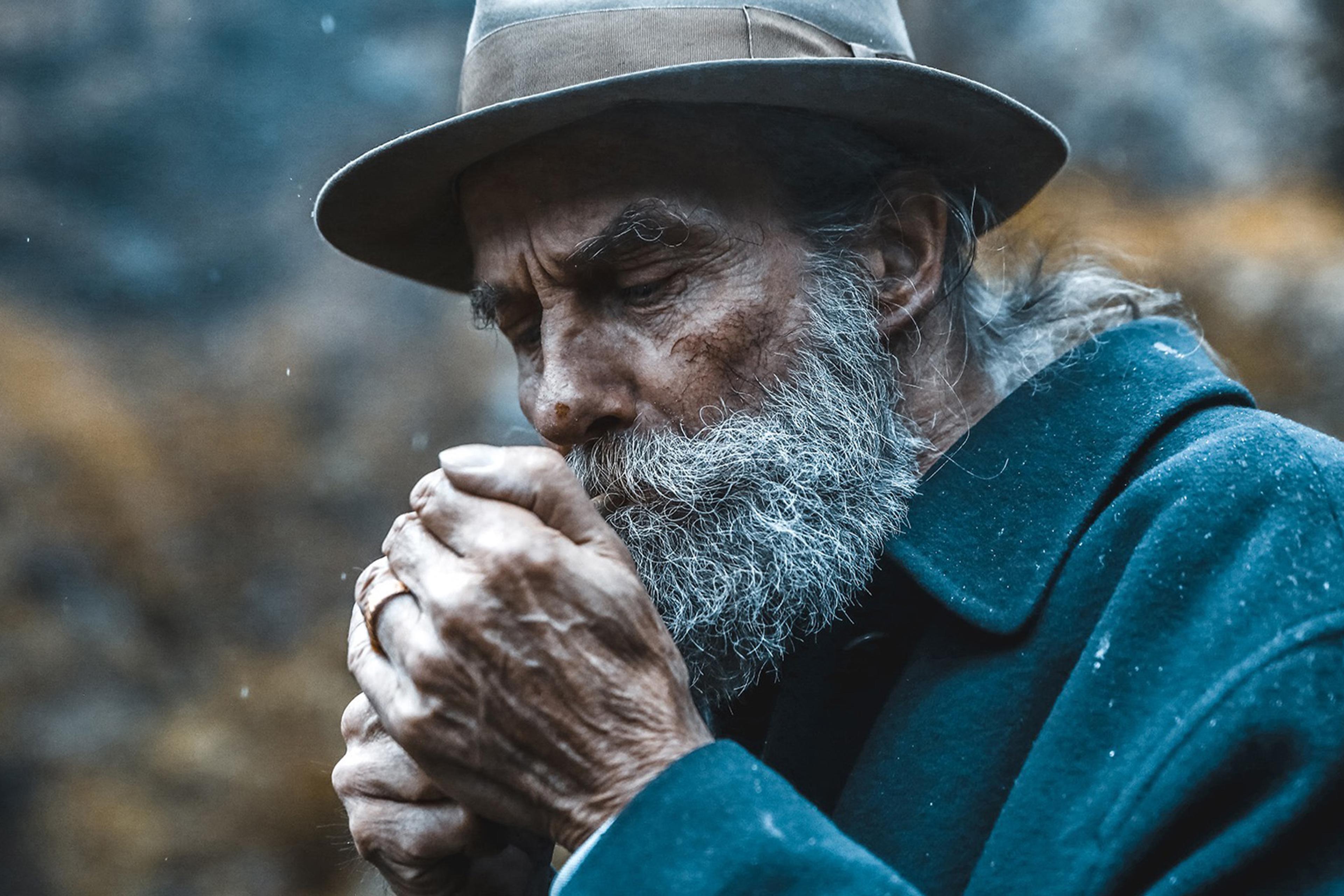 Elderly man in a weathered hat and coat, with a grey beard, lighting a cigarette, set against a blurred outdoor background.