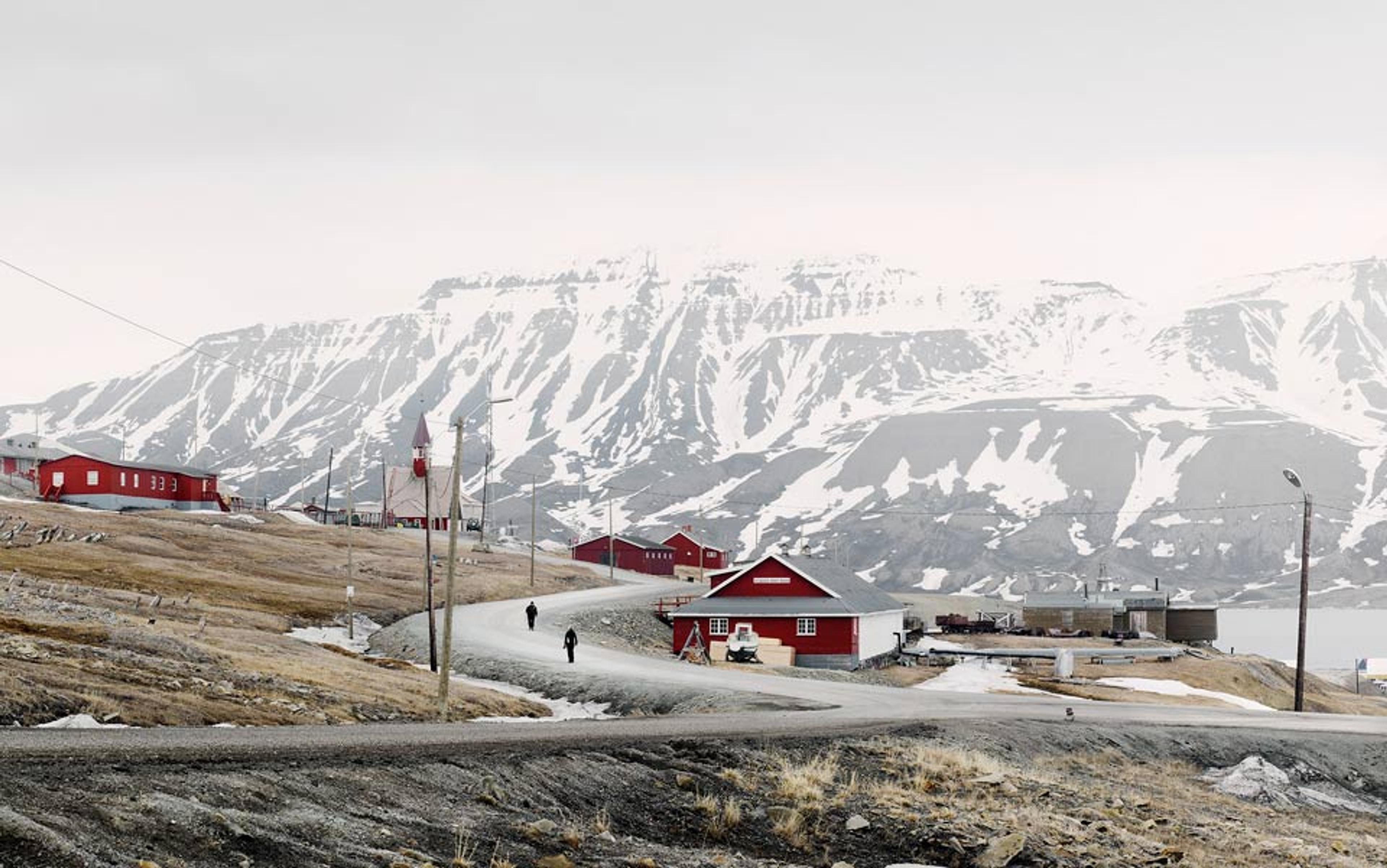 A small settlement with red buildings, snowy mountains in the background, and two people walking on a curving road.
