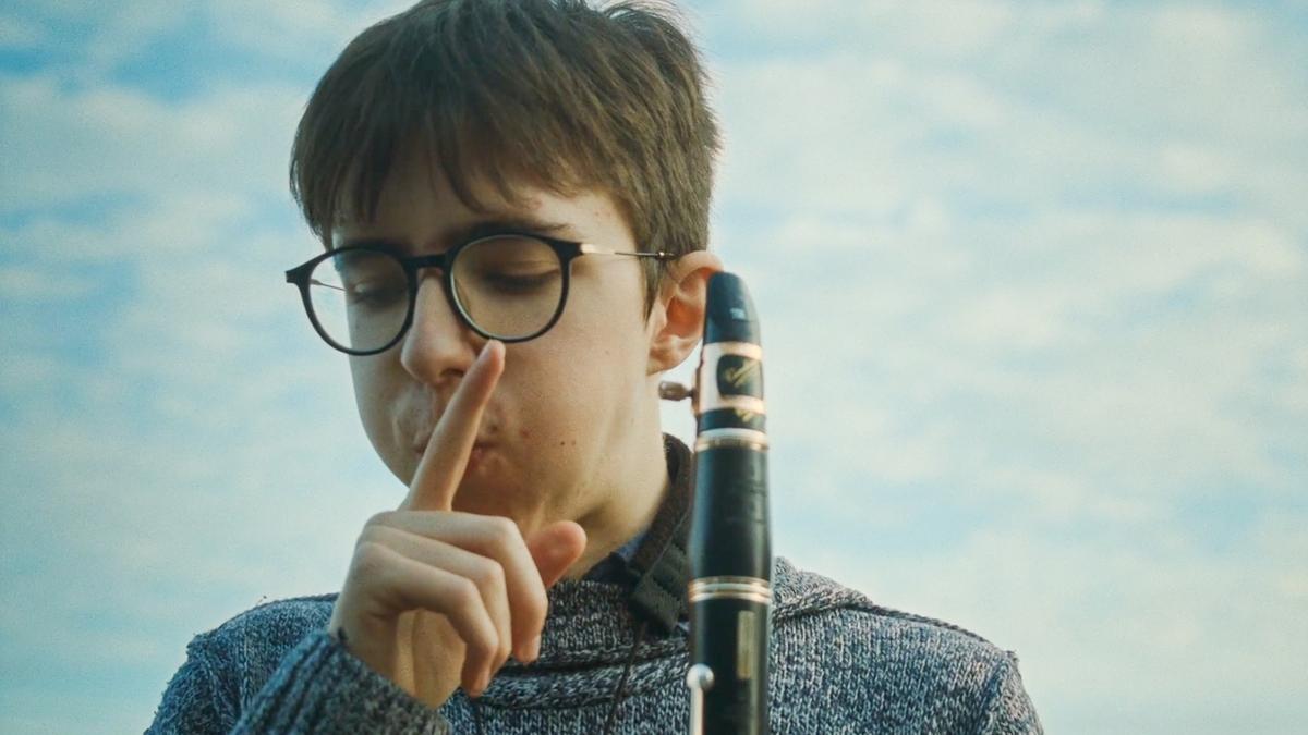 Photo of a person with glasses holding a clarinet, finger to lips in a shushing gesture, blue sky background.