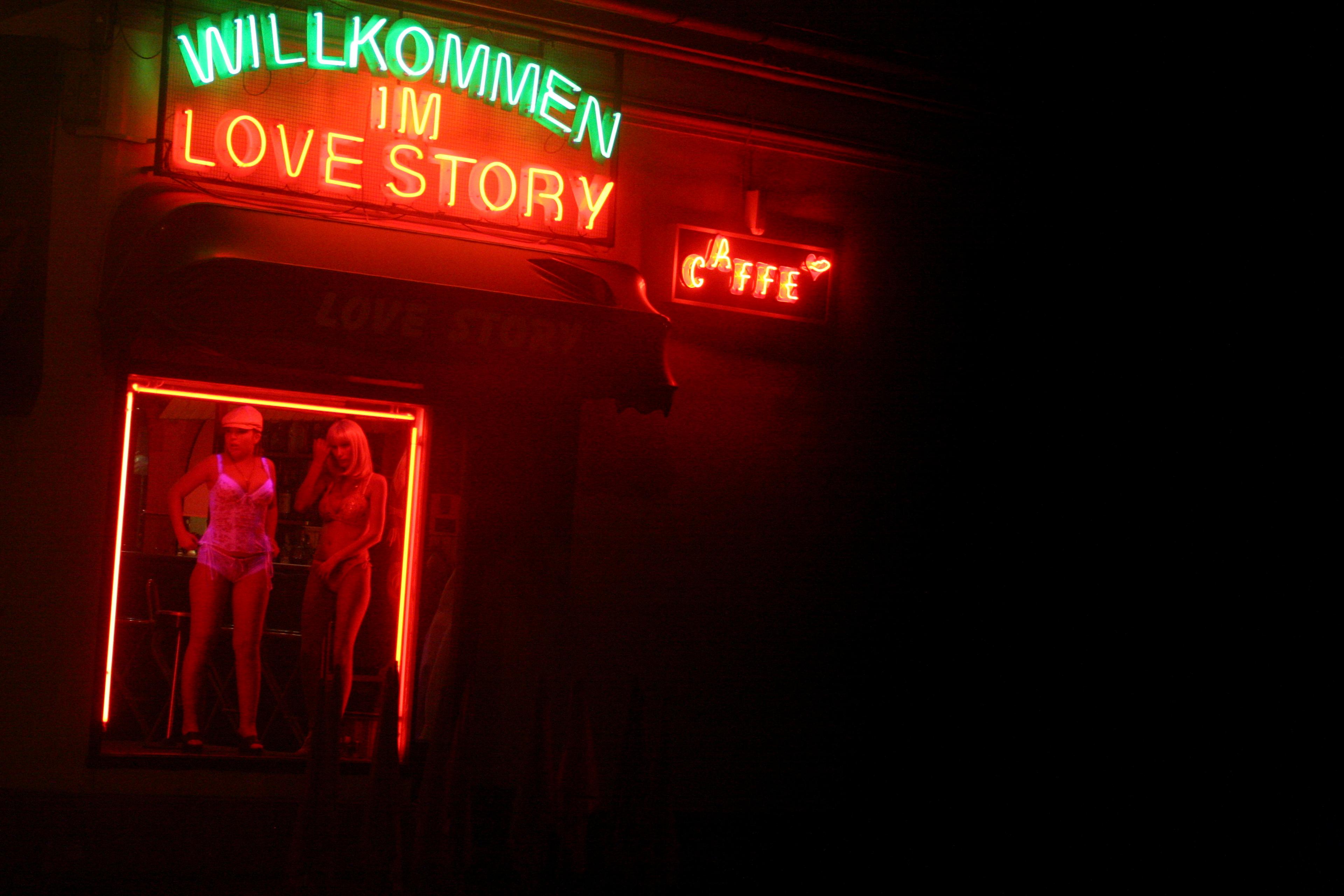 Photo of a neon-lit window display with mannequins, featuring “Willkommen im Love Story” and “Caffe” signs in German.