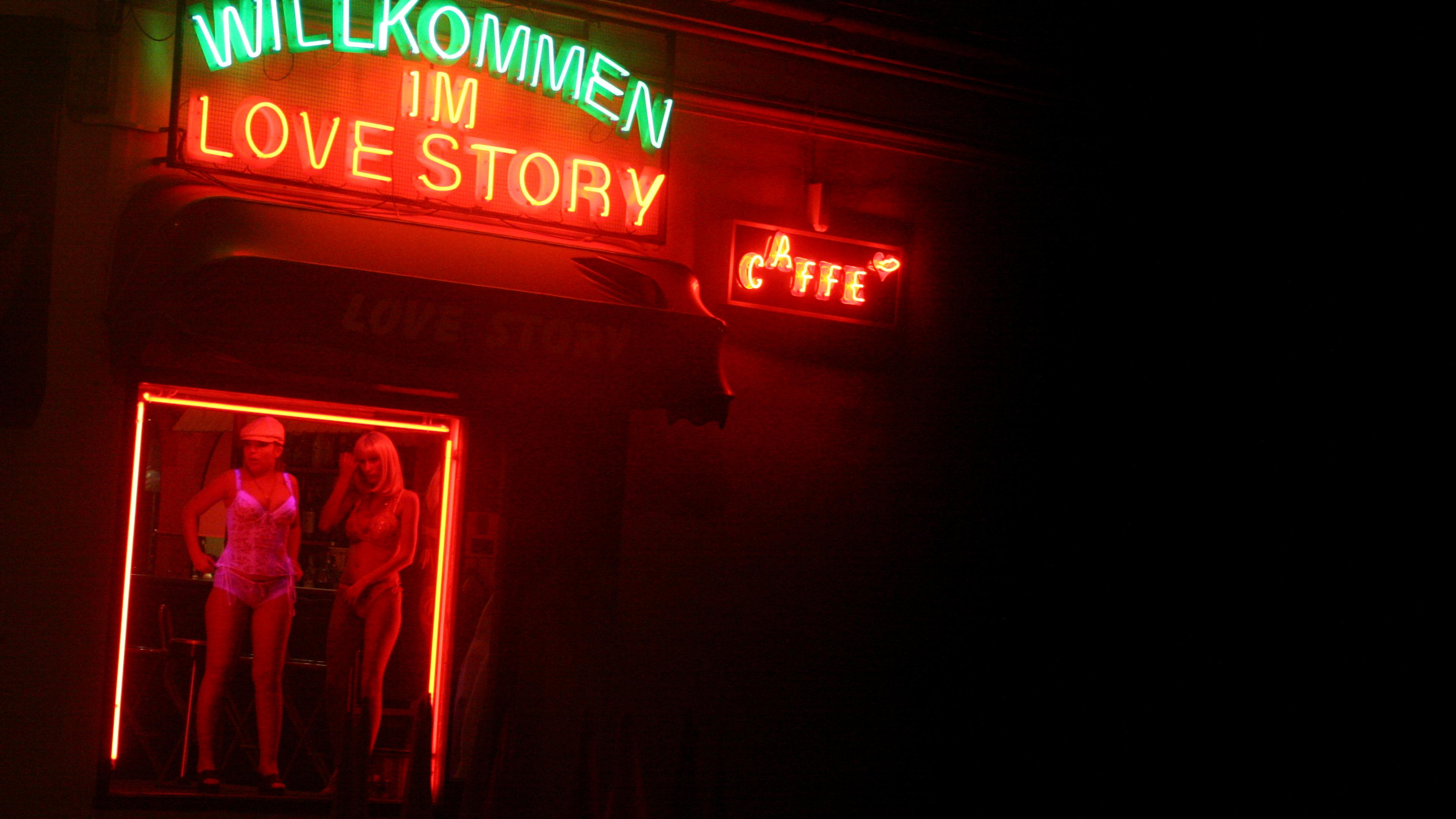 Photo of a neon-lit window display with mannequins, featuring “Willkommen im Love Story” and “Caffe” signs in German.