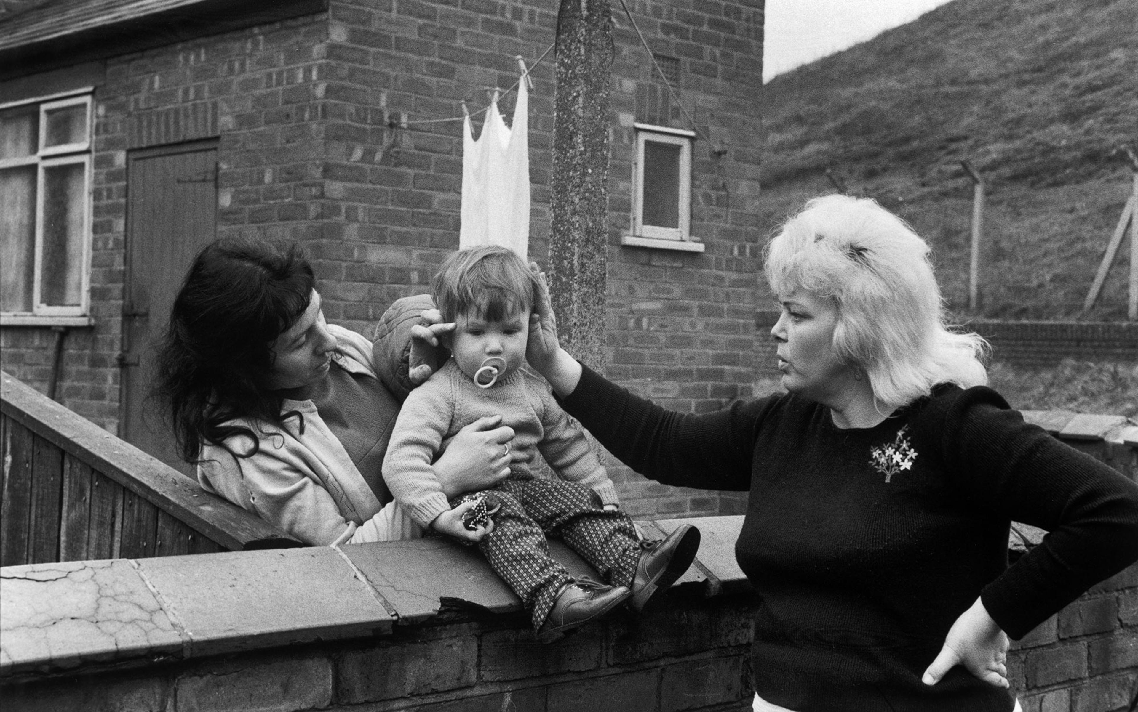 Black-and-white photo of two women preening a toddler who is sitting on a wall and sucking dummy, with a brick house and a washing line in the background.