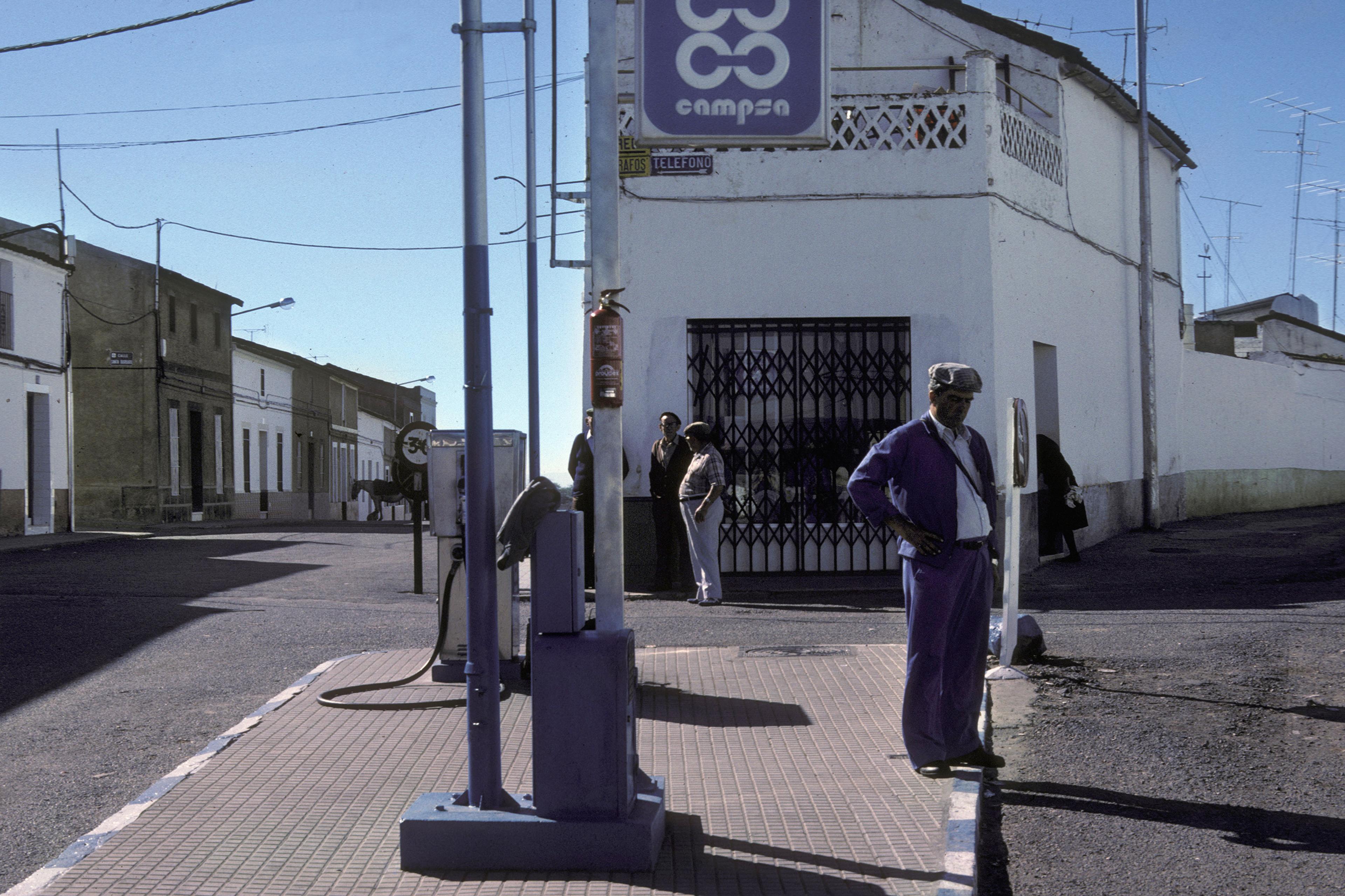 A man standing near a petrol pump on a traffic island between two diverging roads under a clear blue sky.