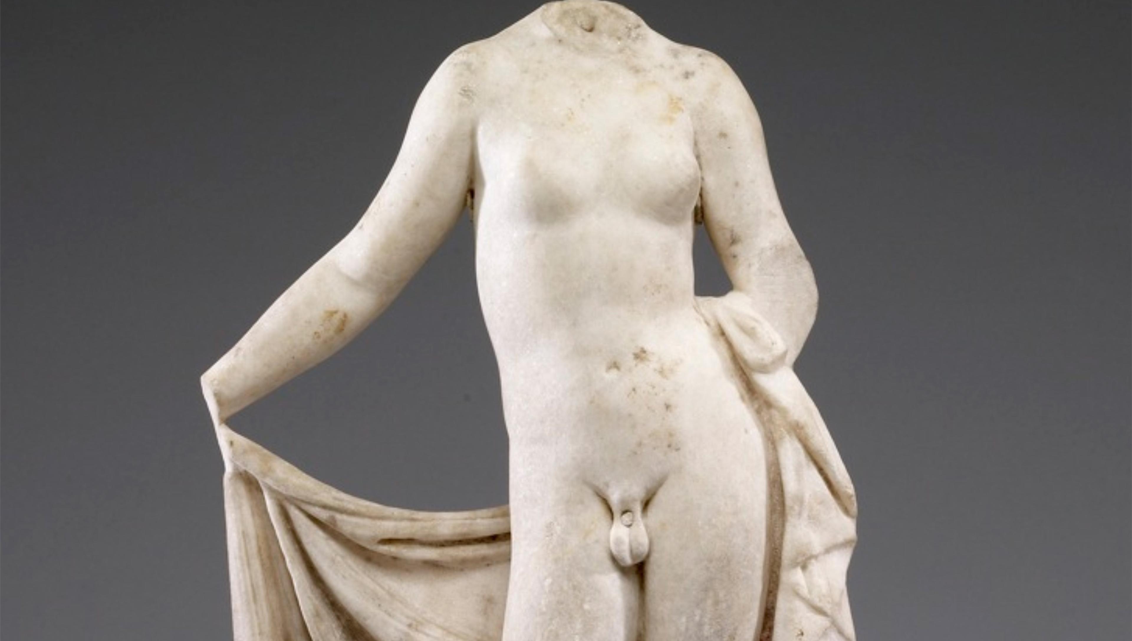 Marble statue of Hermaphroditus, depicting a naked human figure with male and female features, leaning forward slightly and holding a draped cloth in one hand, set against a plain background. The statue lacks a head.
