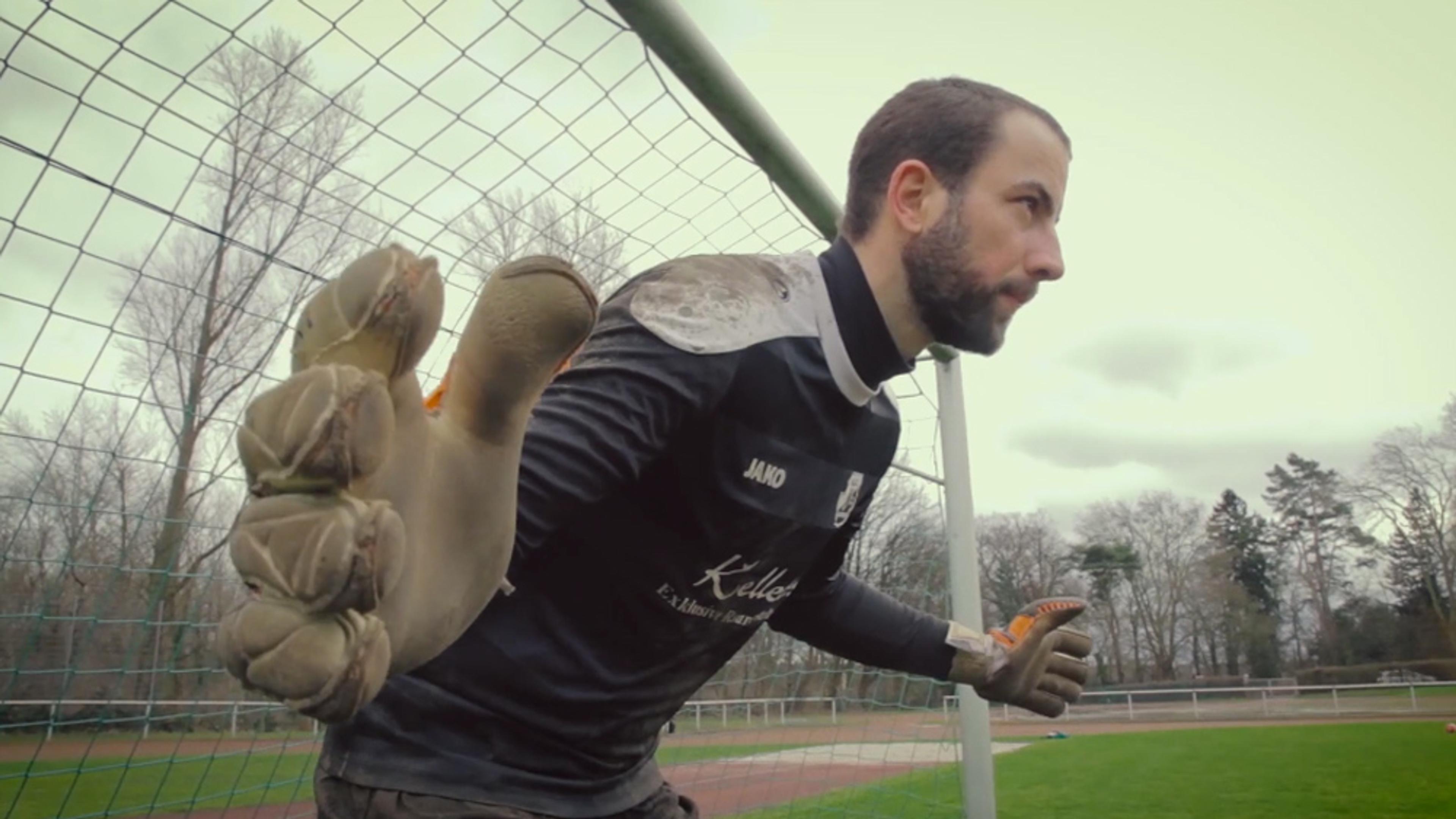 A football goalkeeper in action, wearing gloves, standing in front of the net on an outdoor pitch.