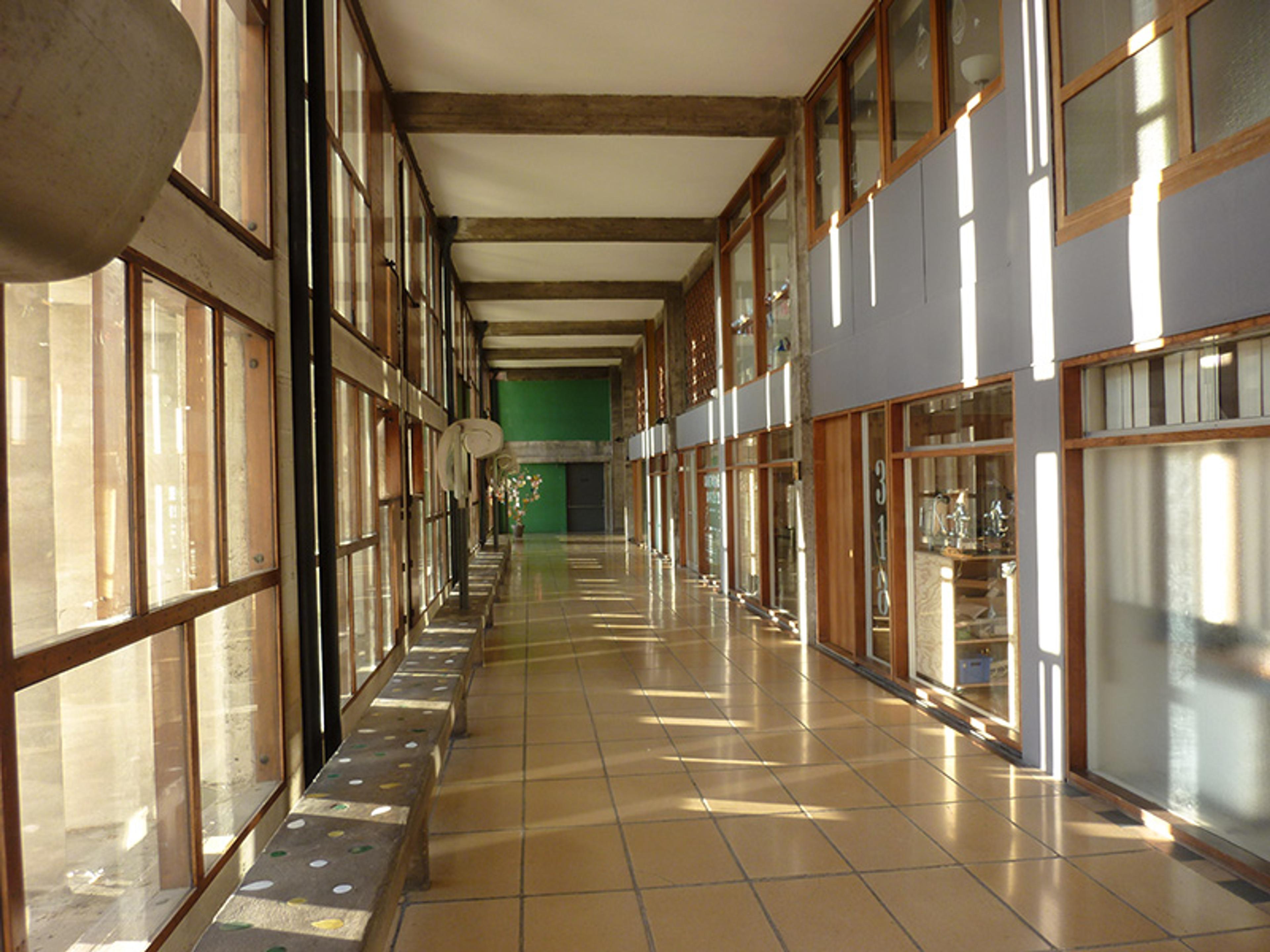 Wide hallway with large windows, concrete benches, wooden beams, tiled floor, and doors on the right, bathed in sunlight.