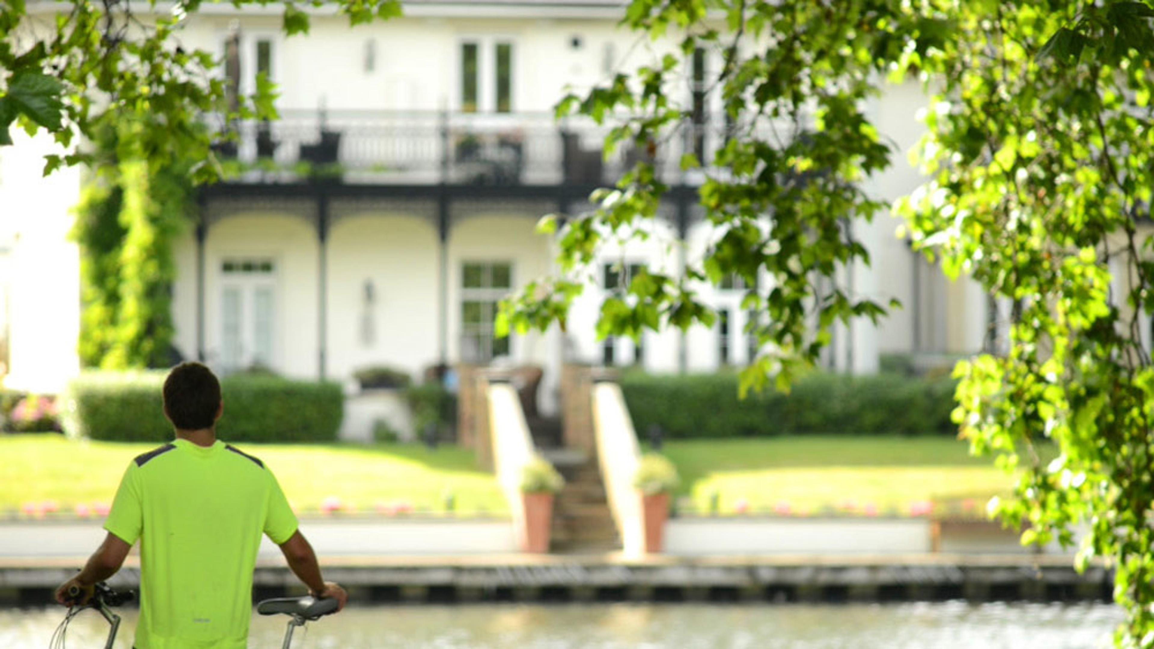 A man in a green shirt with a bicycle looking at a large house with a garden, framed by green tree leaves.