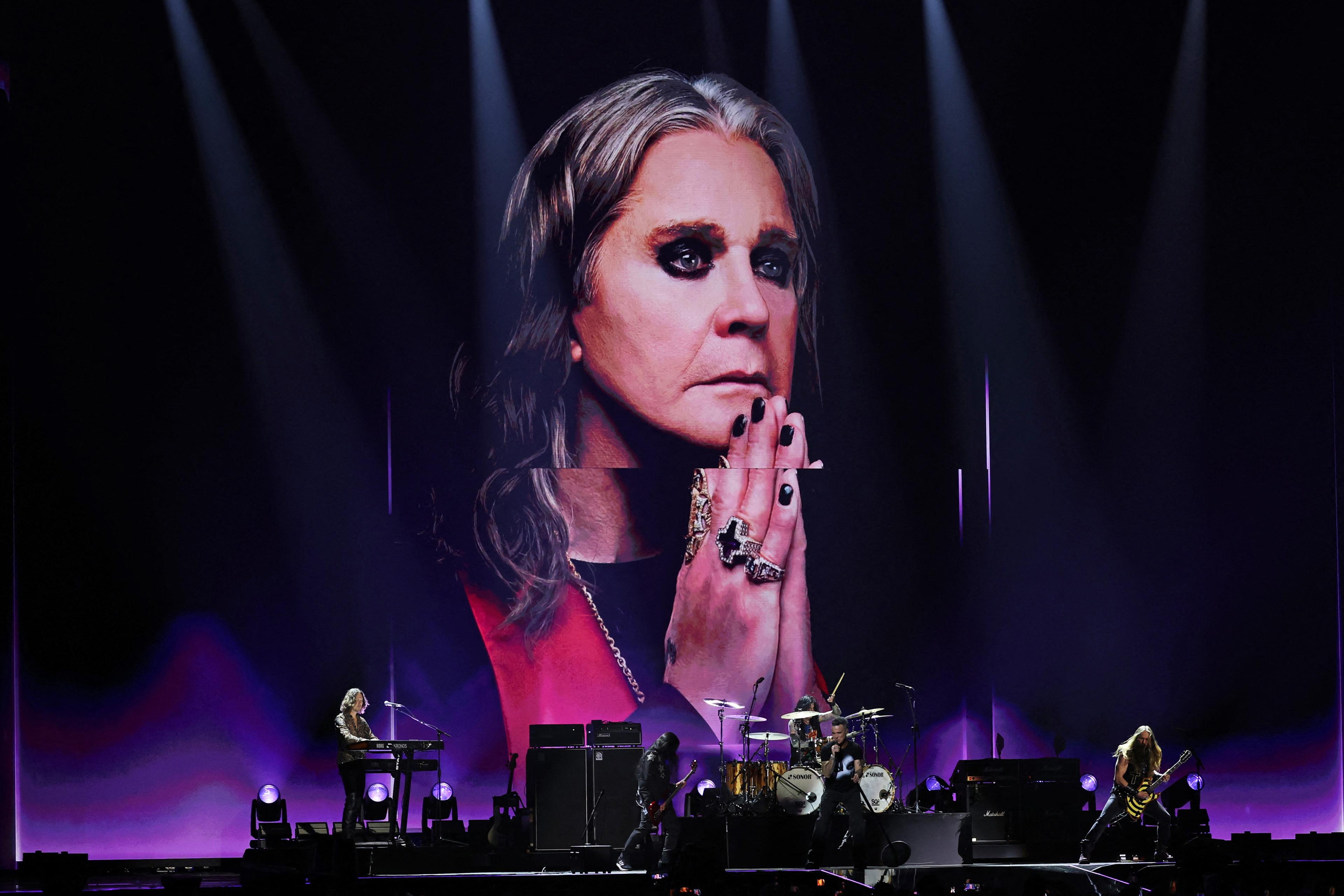 A rock band performing on stage with a large portrait backdrop of a person wearing rings and heavy eye makeup.