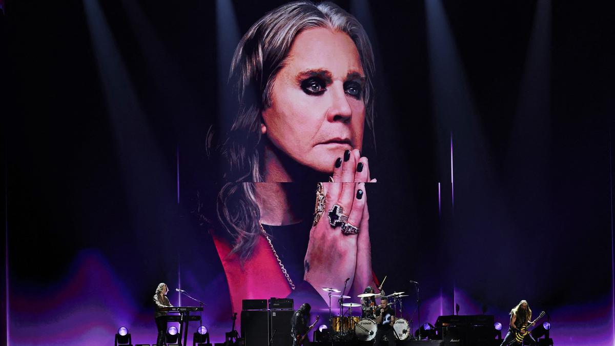 A rock band performing on stage with a large portrait backdrop of a person wearing rings and heavy eye makeup.