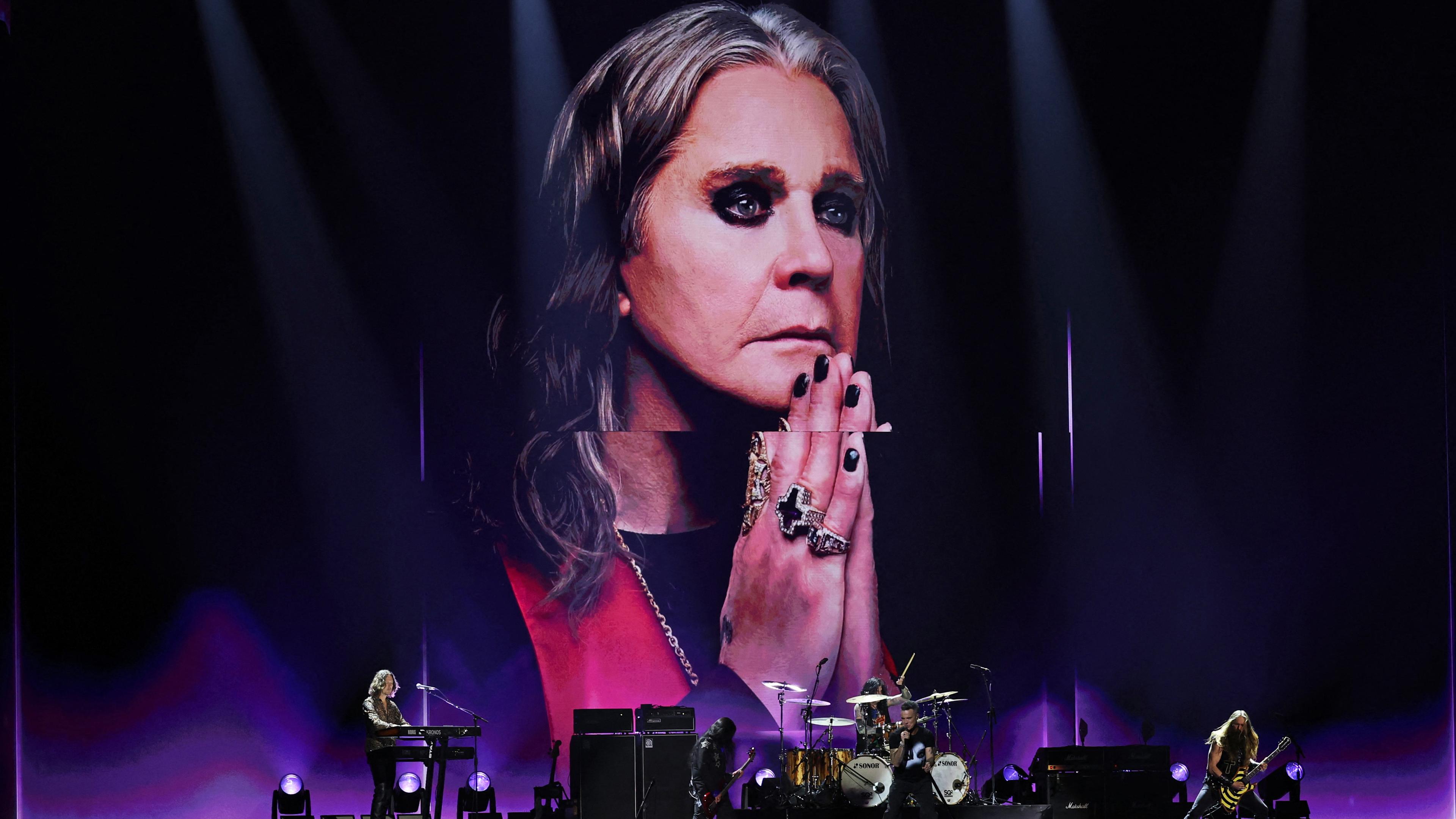 A rock band performing on stage with a large portrait backdrop of a person wearing rings and heavy eye makeup.